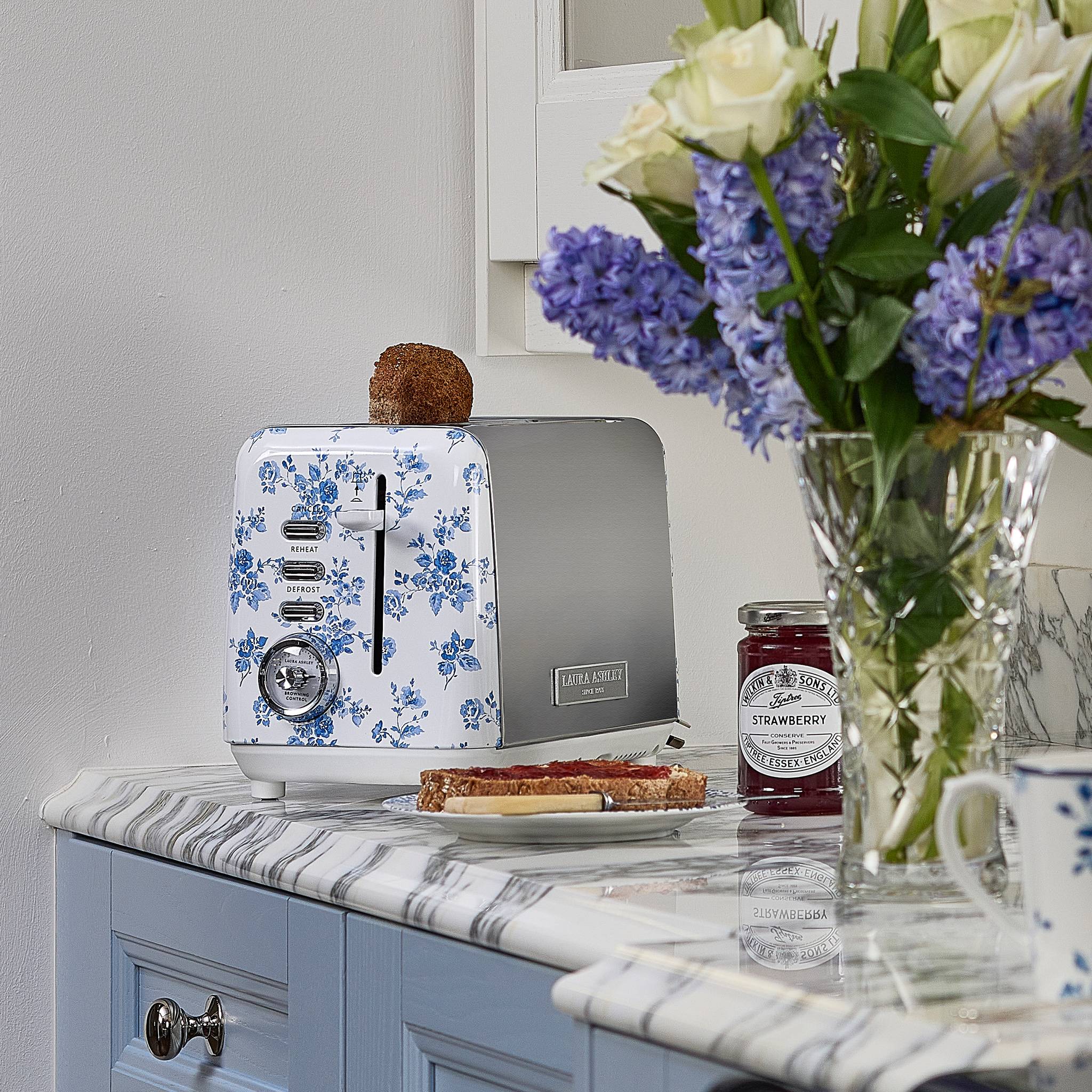 Ein Toaster mit Blumenmuster auf einer Marmorarbeitsplatte mit Toast und Marmelade. Eine Vase mit weißen und violetten Blumen steht in der Nähe in einer hellen Küche.