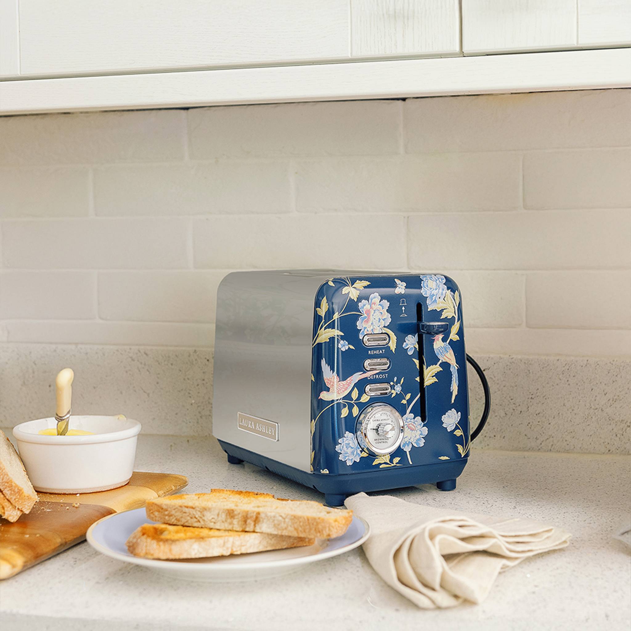 Ein blauer Blumen-Toaster auf einer Küchenarbeitsplatte mit gerösteten Brotscheiben auf einem Teller, einem Schneidebrett und einer Butterdose in der Nähe.