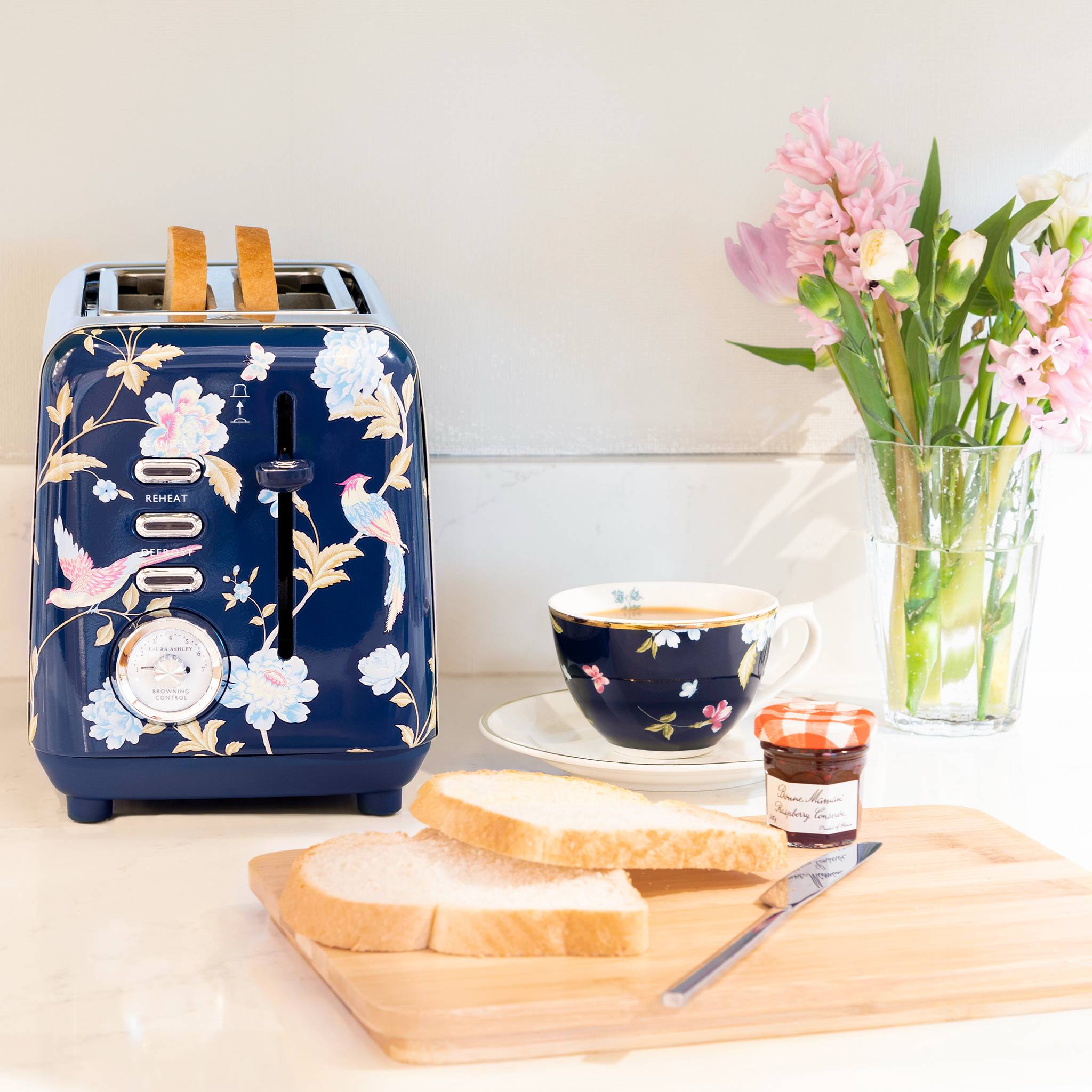 Ein blauer Toaster mit Blumenmuster mit zwei Brotscheiben. Daneben eine Tasse mit passendem Design, Blumen und ein Glas Marmelade auf einem Holzbrett.