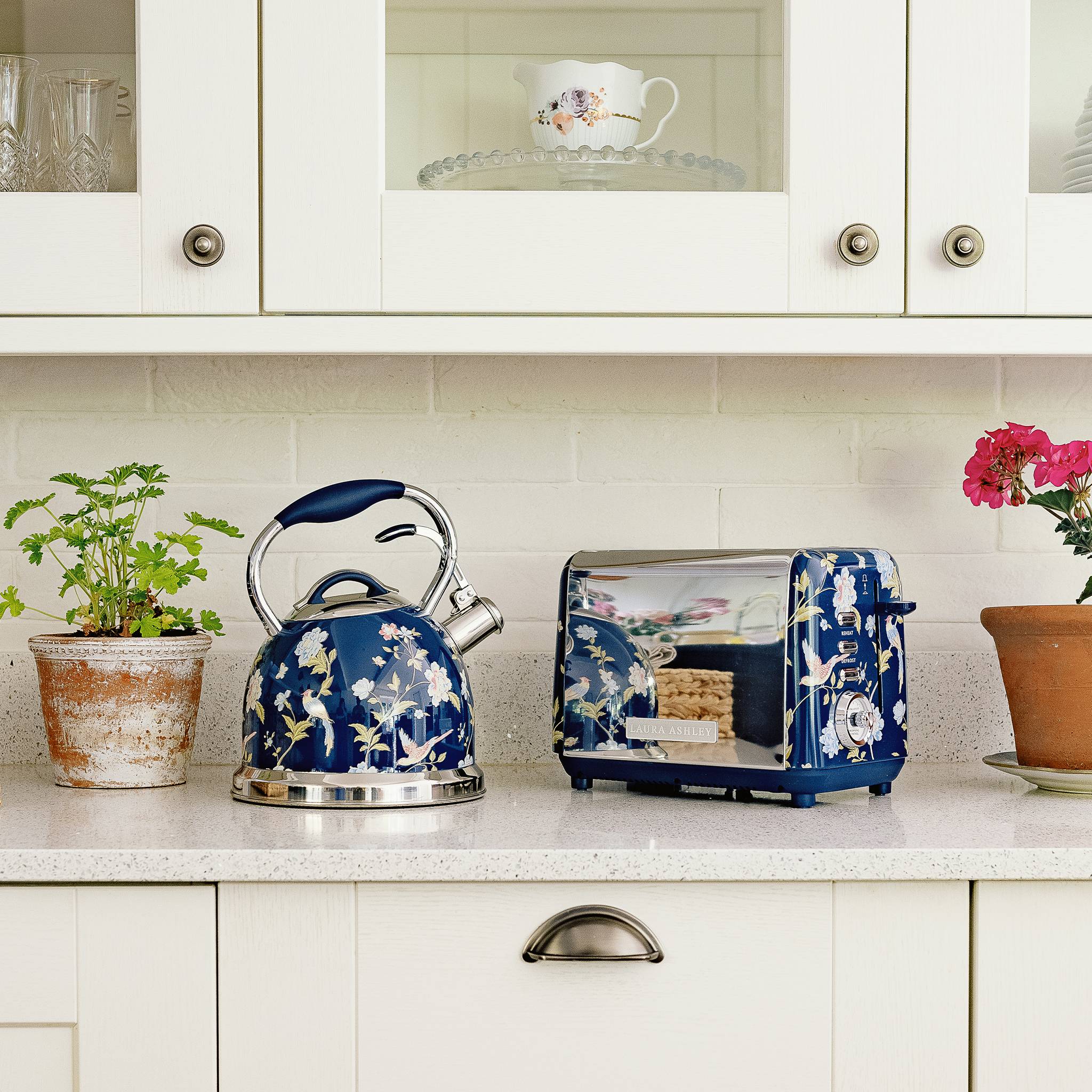 Eine Küchenarbeitsplatte mit einem dekorativen blauen Wasserkessel und einem Toaster mit Blumenmotiven. In der Nähe befinden sich Topfpflanzen und eine rosa Blumenvase.