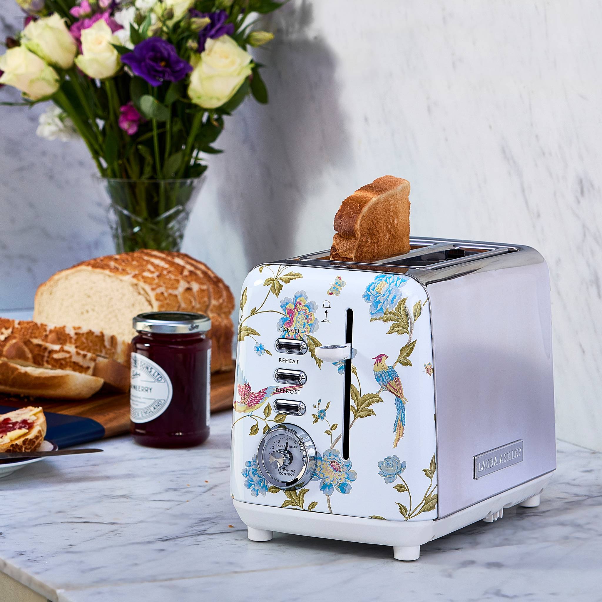 Ein dekorativer Toaster mit Blumenmotiven, der zwei Scheiben getoastetes Brot hält, steht auf einer Küchentheke neben einem Brotlaib und einem Glas Marmelade. Eine Vase mit bunten Blumen steht im Hintergrund.