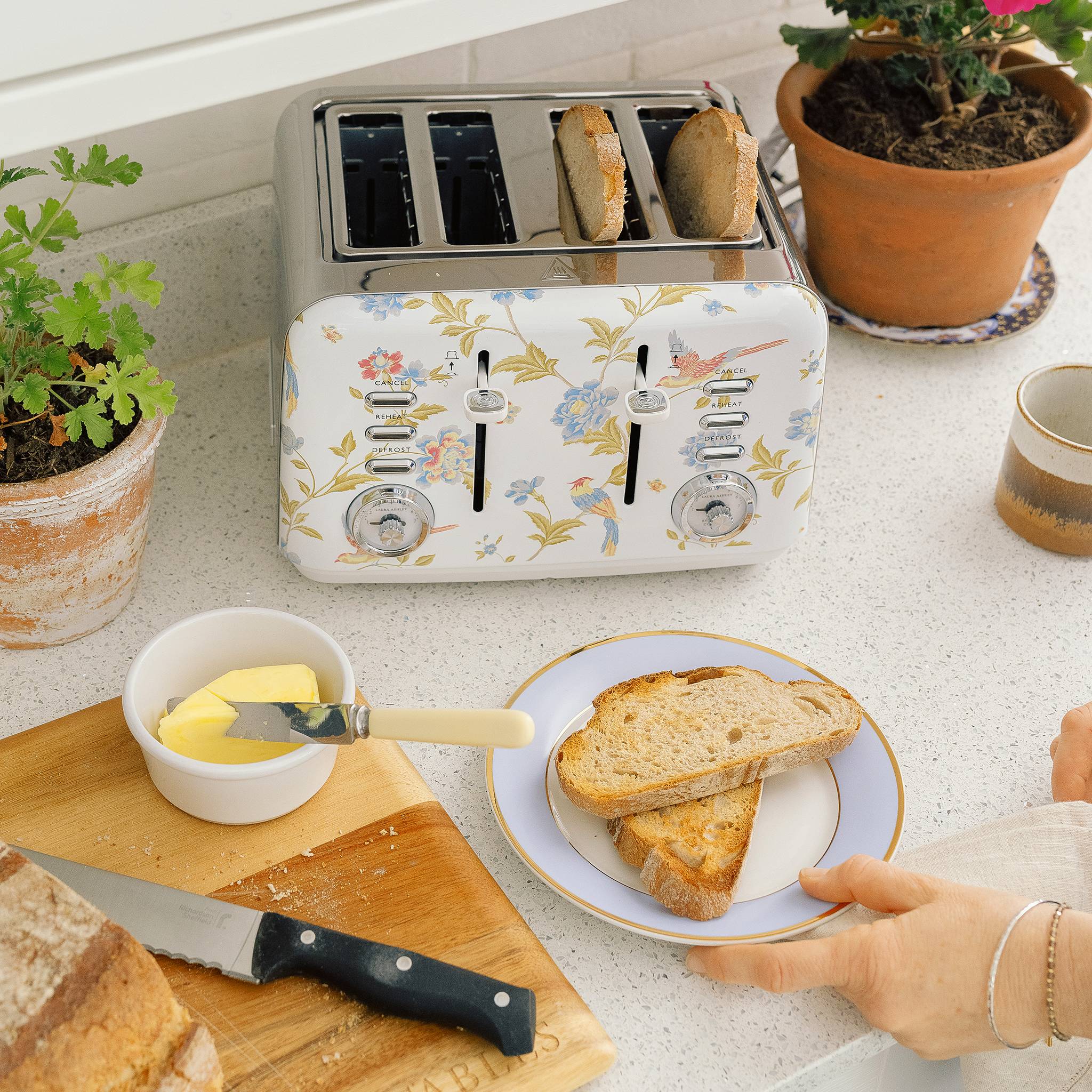 Eine Person bereitet Frühstück mit getostetem Brot, Butter und einem Retro-Toaster mit floralem Muster auf einer Küchenablage neben Topfpflanzen zu.