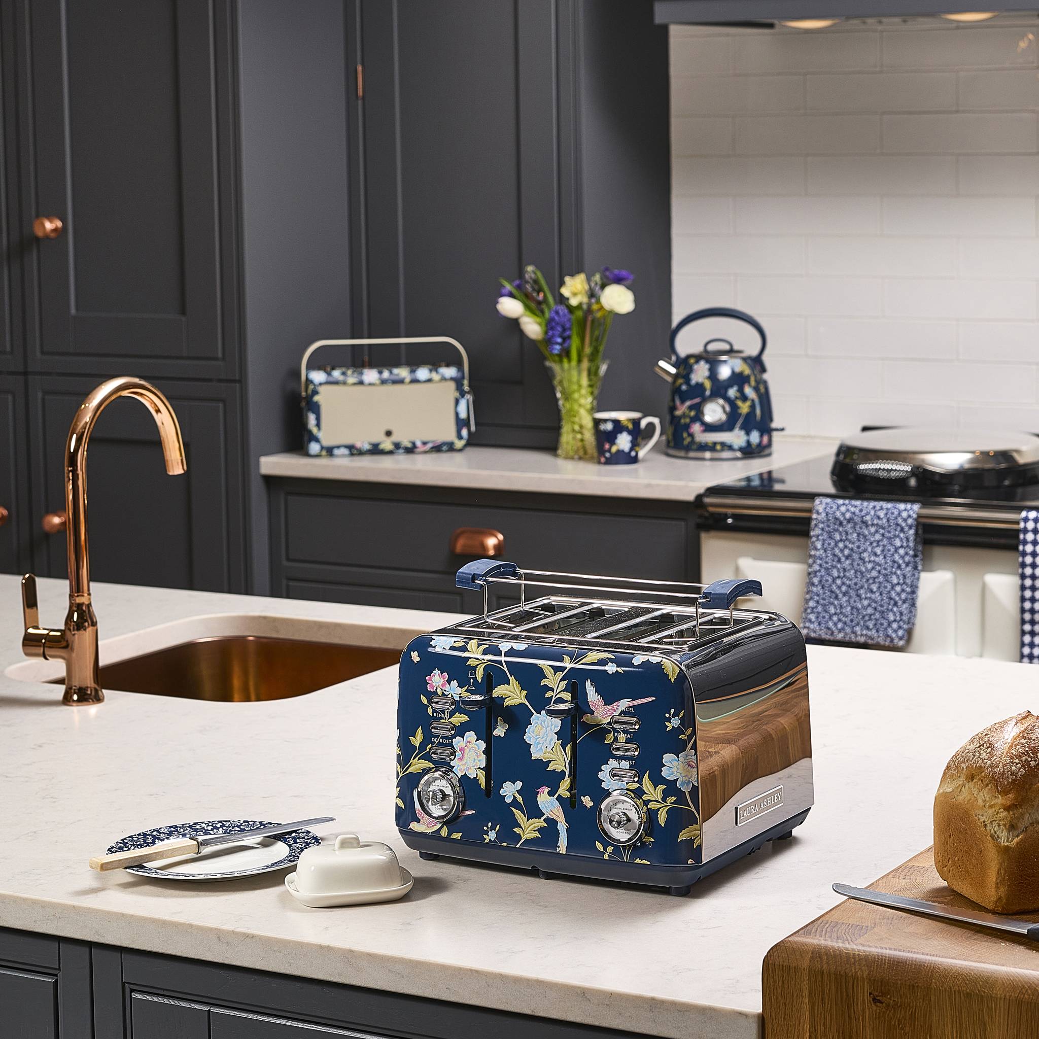 Eine moderne Küche mit blauem Blumenmuster-Geräten, einschließlich eines Toasters und eines Wasserkochers, auf einer Arbeitsplatte mit einer kupferfarbenen Armatur und einer Vase mit Blumen.