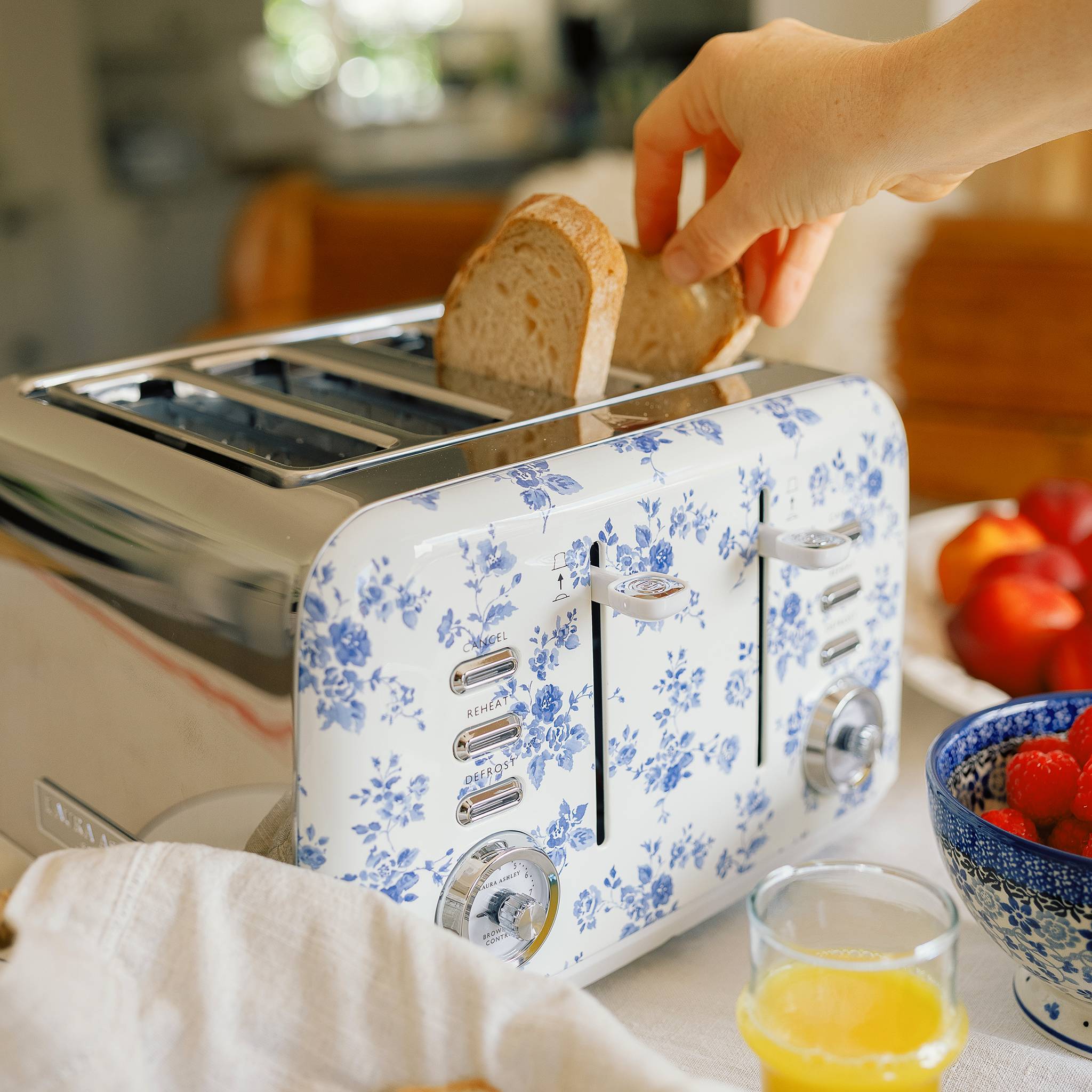 Eine Person legt Brot in einen Toaster mit blauen Blumenmustern. Eine Schale mit Obst und ein Glas Orangensaft stehen in der Nähe auf dem Tisch.