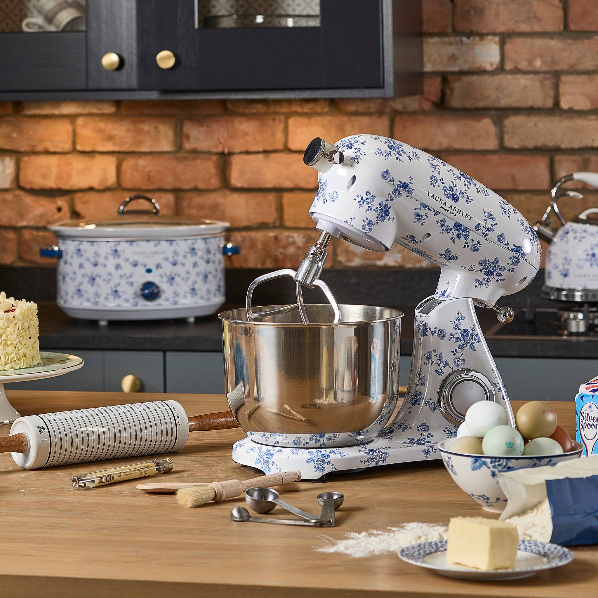 Ein blauer Standmixer mit Blumenmuster auf einer Küchentheke mit Backutensilien, Butter, Mehl und Kuchen vor einer rustikalen Ziegelmauer.