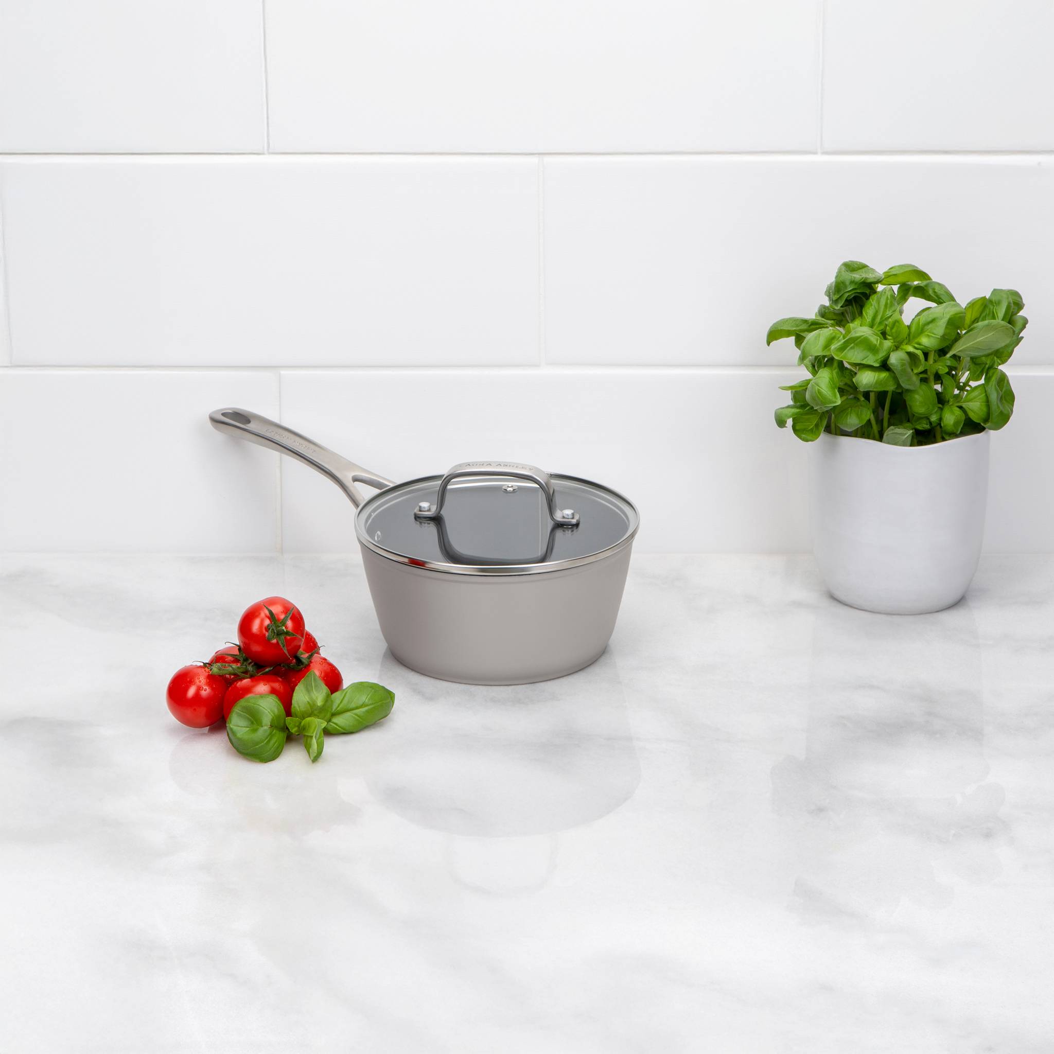 Ein Kochtopf mit Deckel auf einer Küchenarbeitsplatte neben frischen Tomaten, Paprika und einer Topfpflanze mit Basilikum an einer weiß gefliesten Wand.