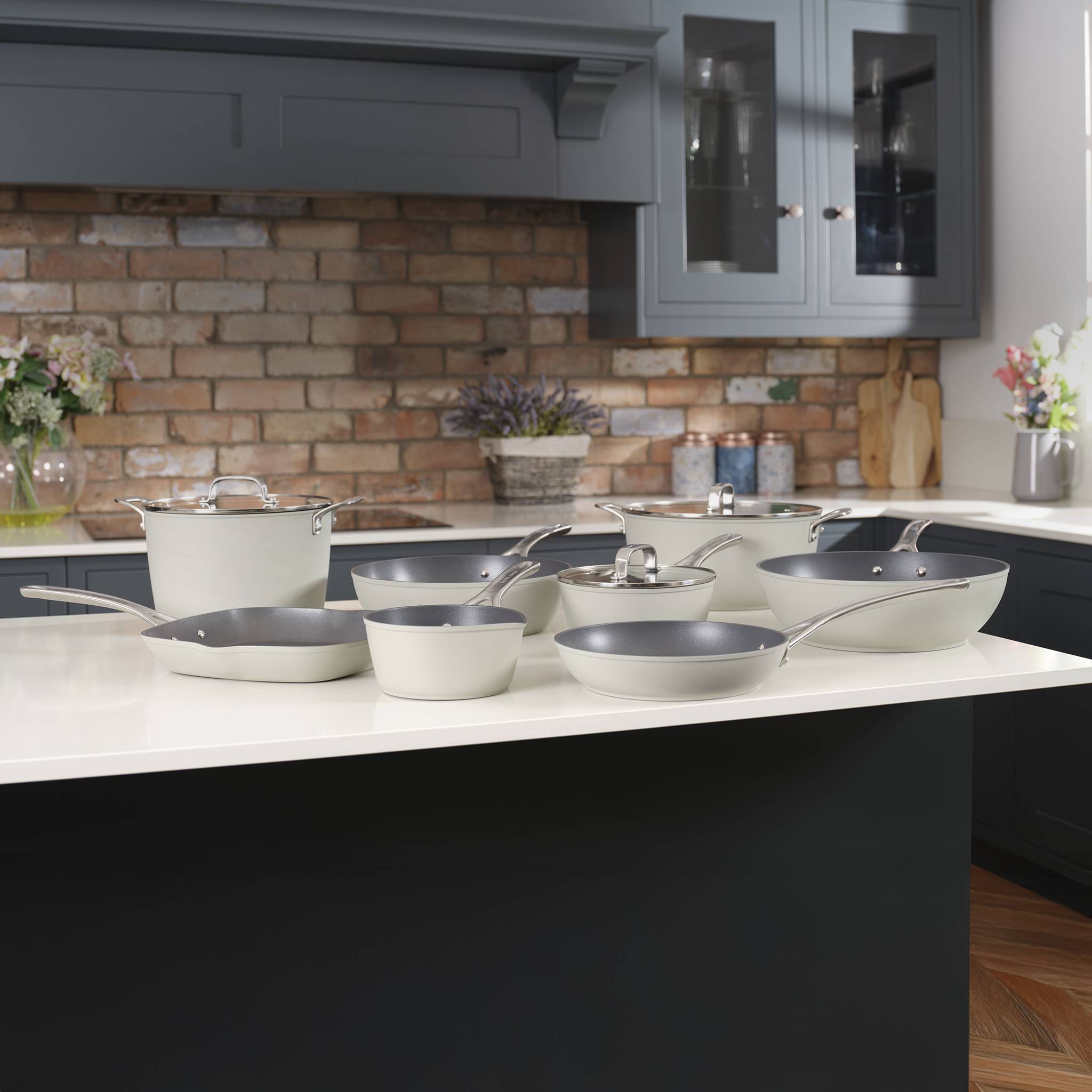 Ein Sortiment verschiedener Kochgeschirre, bestehend aus Töpfen und Pfannen, ist auf einer Küchentheke angeordnet. Der Hintergrund zeigt eine Backsteinwand und Schränke.