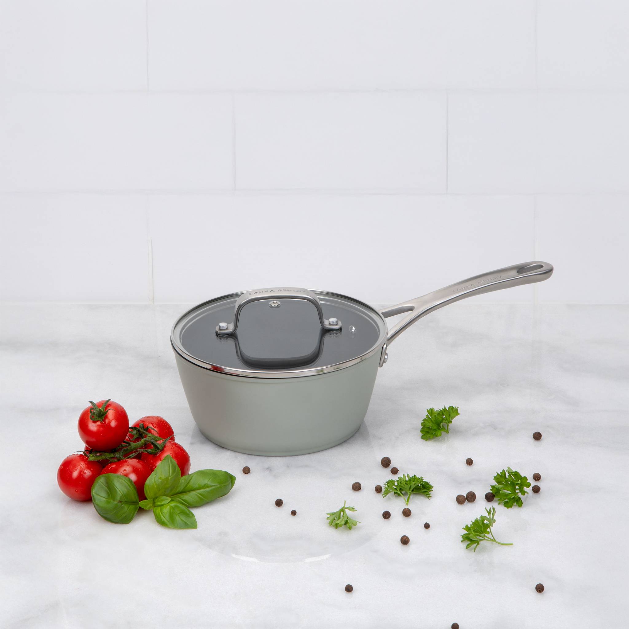 Ein Kochtopf mit Deckel auf einer Marmorarbeitsfläche, umgeben von frischen Tomaten, Basilikumblättern, Petersilie und Pfefferkörnern vor einer gekachelten Wand.