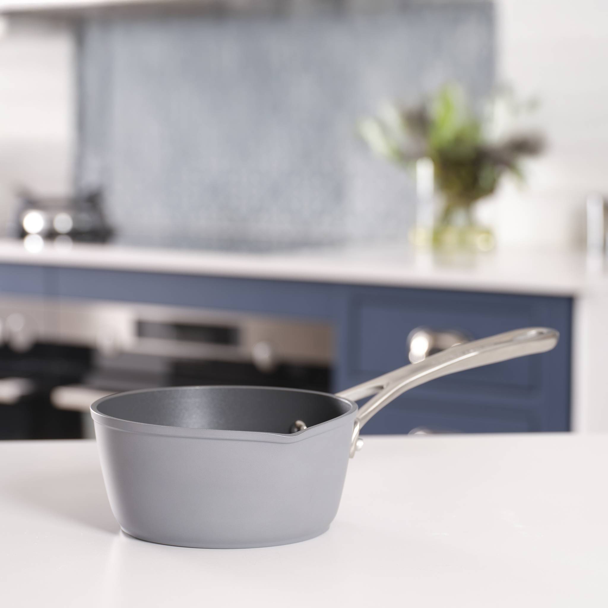 Eine graue Pfanne mit metallischem Griff sitzt auf einer weißen Küchenarbeitsplatte. Der unscharfe Hintergrund zeigt eine moderne Kücheneinrichtung.