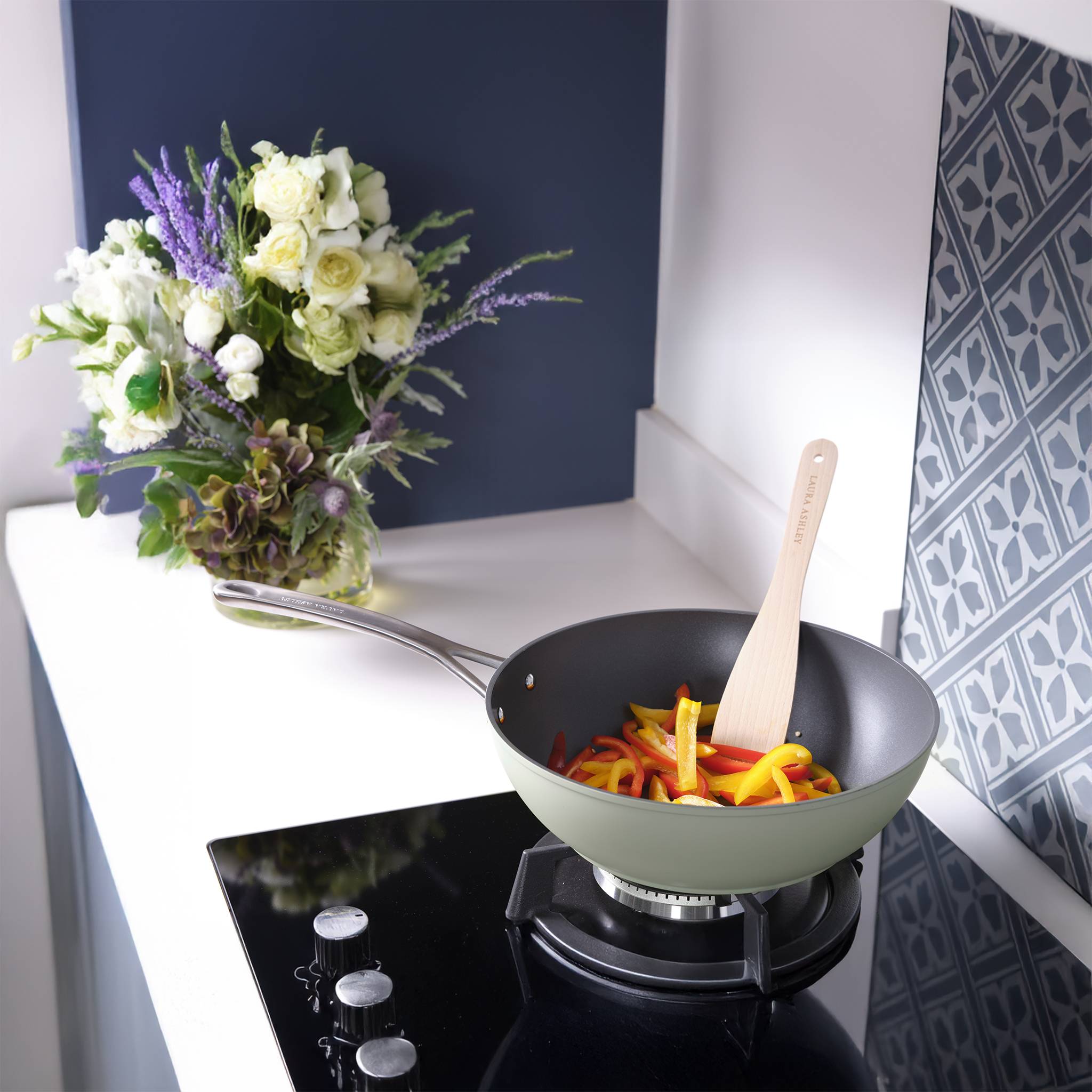 Ein Topf mit geschnittenen Paprikaschoten, die auf einem Gasherd kochen, umgeben von dekorativen Blumen auf einer Arbeitsplatte.