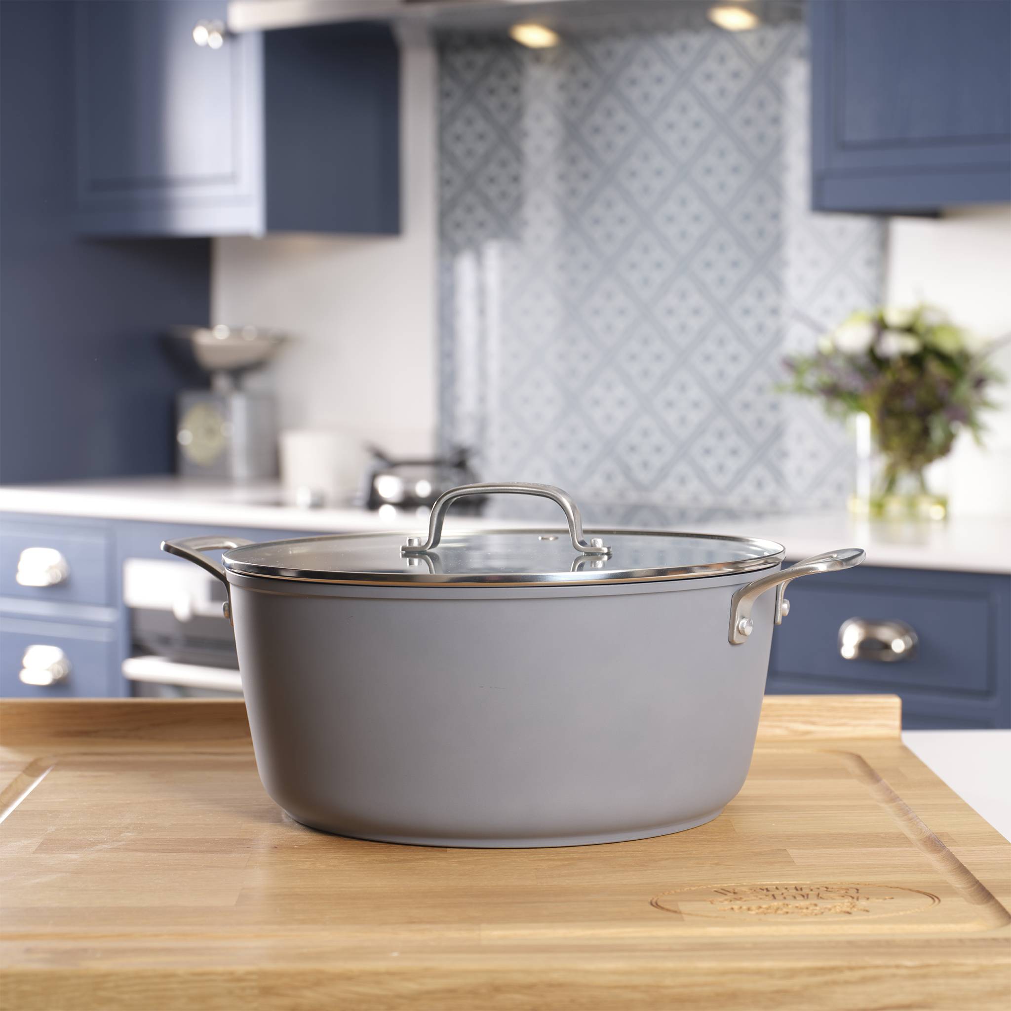 Ein grauer Kochtopf mit Glasdeckel steht auf einer hölzernen Arbeitsplatte in einer modernen Küche mit blauen Schränken und dekorativer Fliesenwandverkleidung.