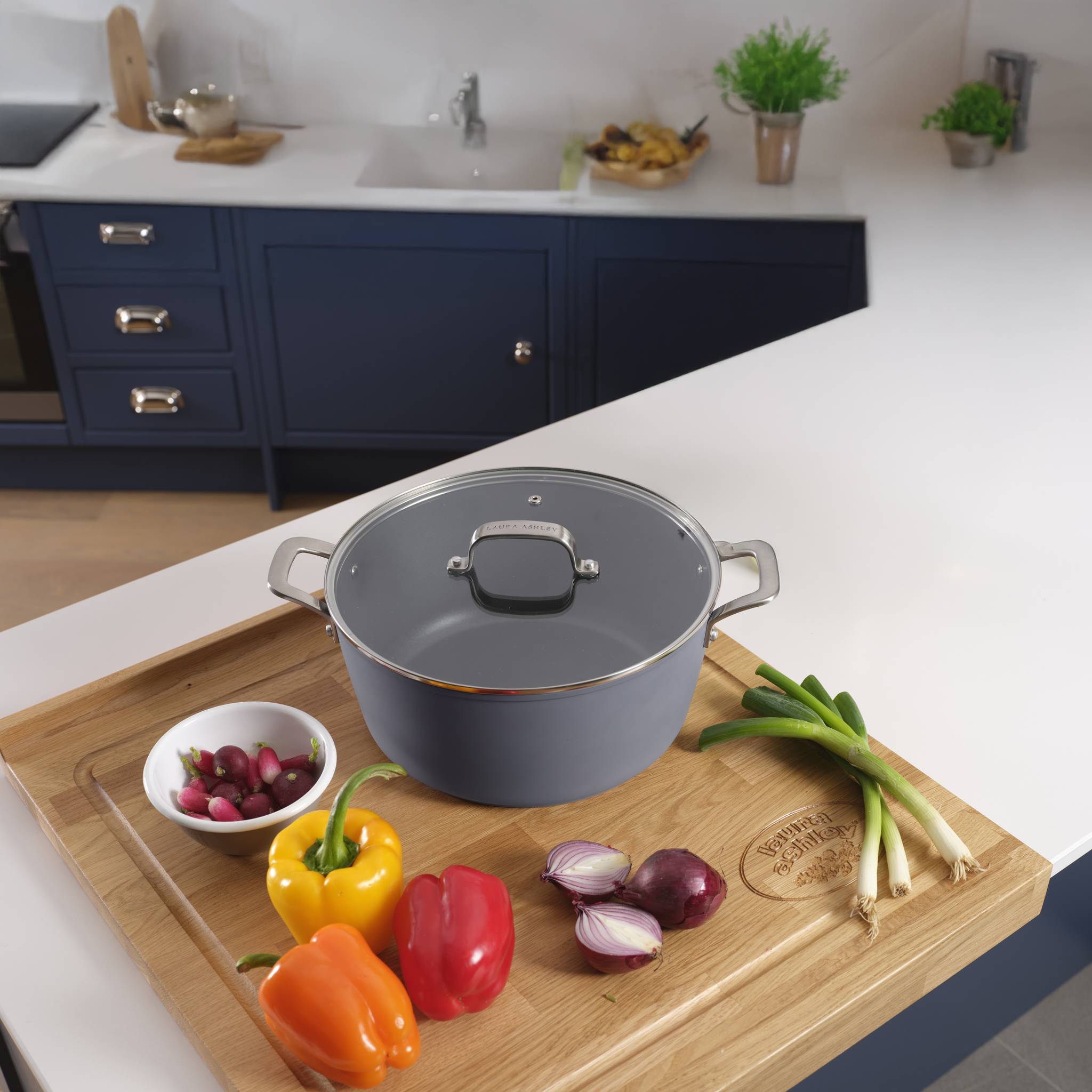 Eine Küchenszenerie mit einem großen Topf auf einem Holzbrett, umgeben von farbenfrohen Paprikaschoten, Zwiebeln und Radieschen.