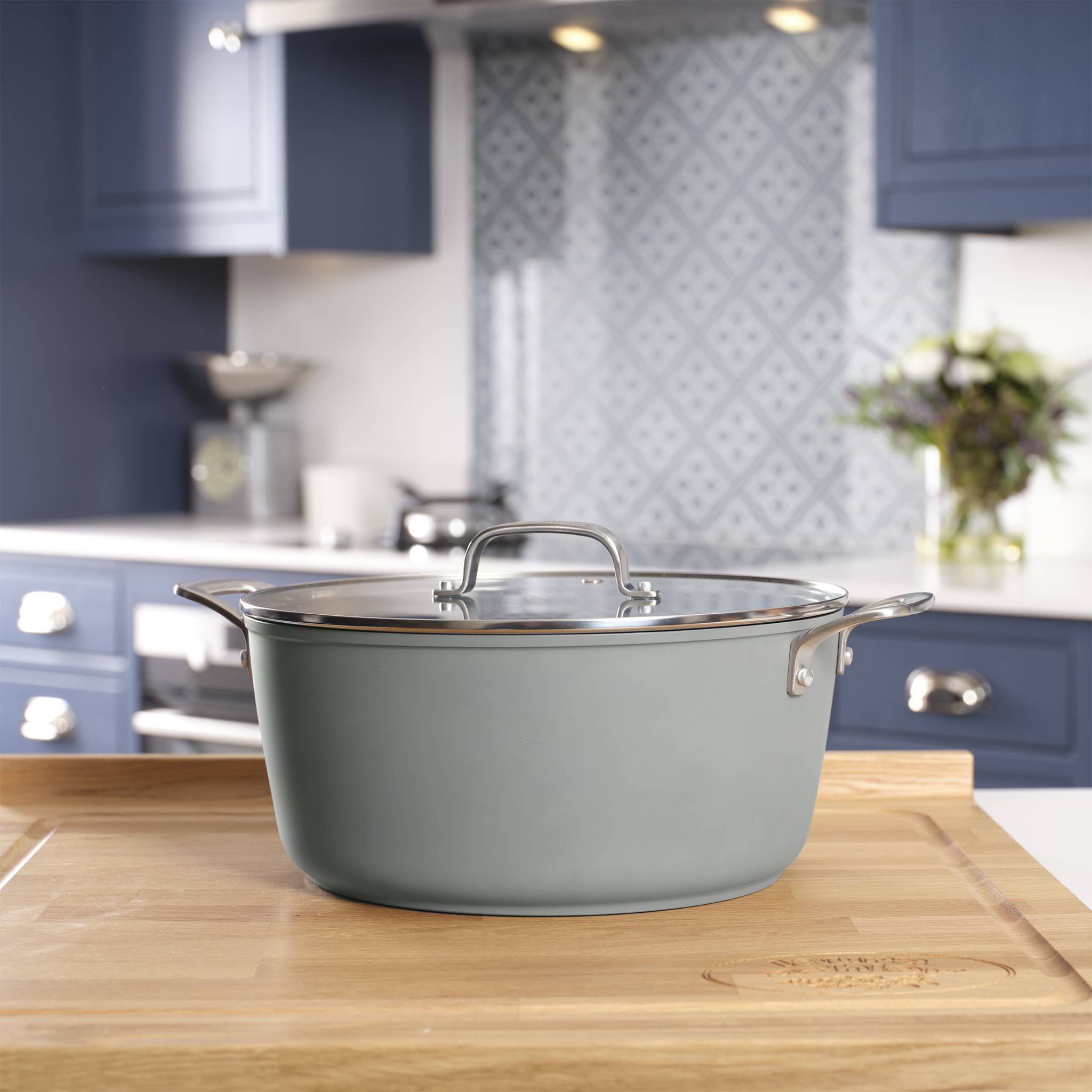 Ein großer, blauer Kochtopf mit einem Glasdeckel sitzt auf einer Holzarbeitsplatte in einer modernen Küche mit blauen Schränken und gemusterter Rückwand.