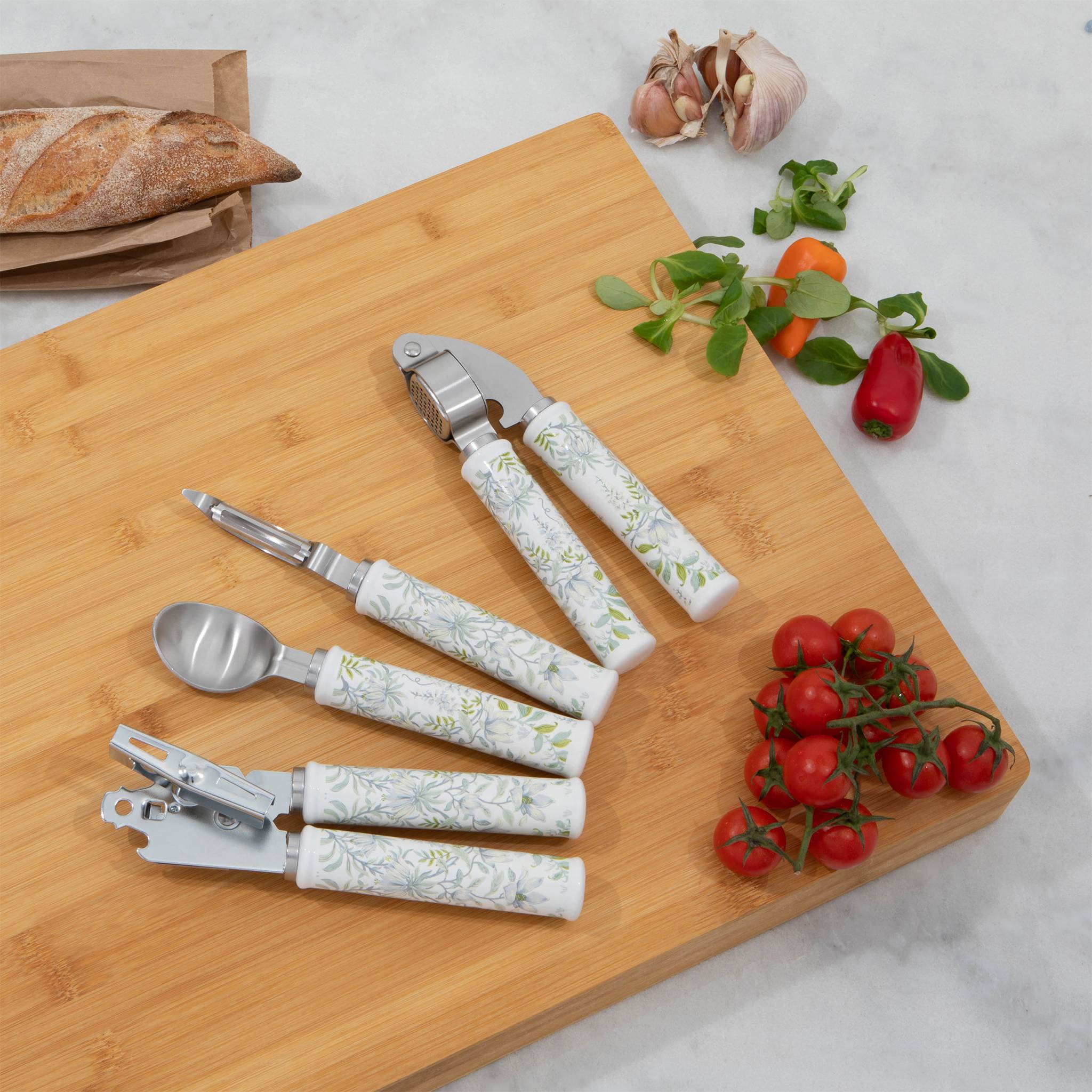 Ein Schneidebrett aus Bambus mit einer Knoblauchpresse, einem Schäler, einem Dosenöffner und einem Utensil mit floralen Griffen. Kirschtomaten und Kräuter sind in der Nähe.