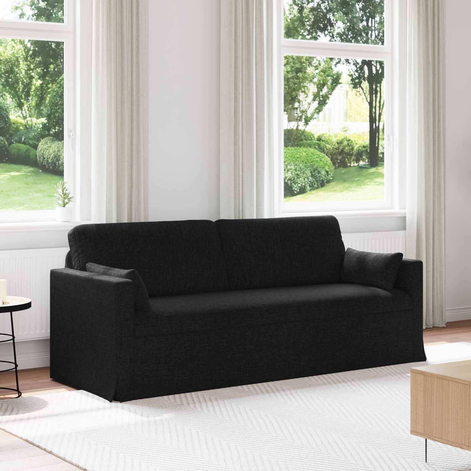 Ein schwarzes Sofa steht in einem hellen Wohnzimmer mit großen Fenstern und Grünflächen, die von außen sichtbar sind. Ein heller Teppich und ein Holztisch vervollständigen die Einrichtung.