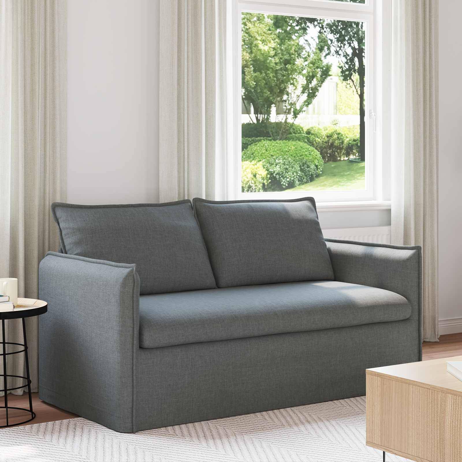 Ein modernes Wohnzimmer mit grauem Sofa, hellem Holz-Couchtisch und großen Fenstern, die das Grün draußen zeigen. Der Raum ist hell und luftig.