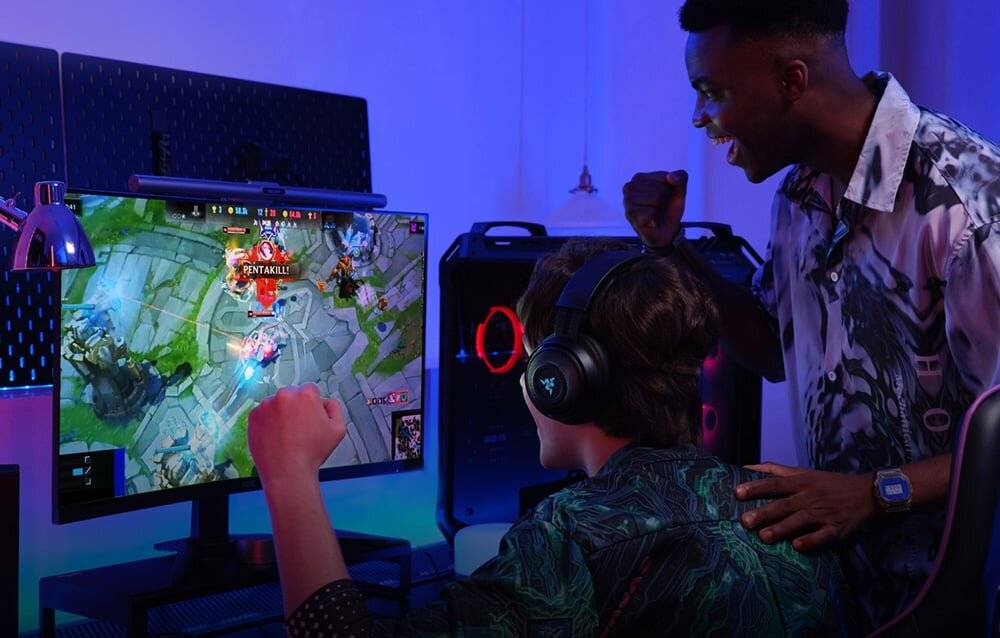 Zwei Personen beobachten aufgeregt ein Videospiel auf einem Monitor in einem dunklen Zimmer, eine trägt Kopfhörer und hält einen Game-Controller, während beide einen Sieg feiern.