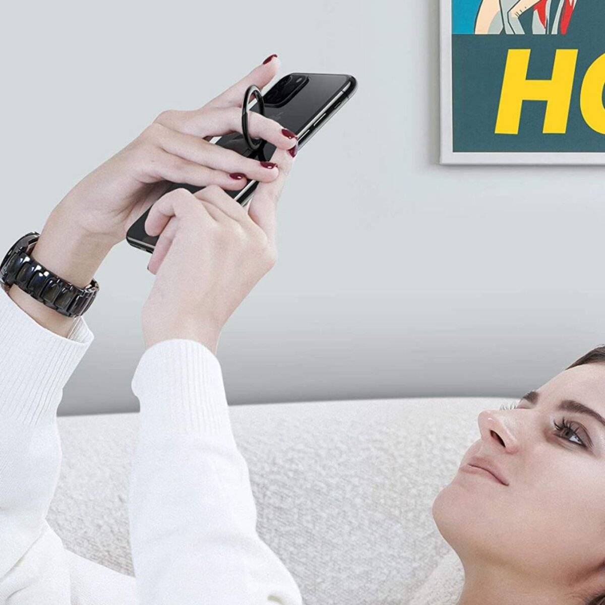 Eine Person, die liegt, nutzt ein Smartphone mit angeschlossenem Ladekabel. Ein bundes Kunstwerk in einem Rahmen ist teilweise an der Wand sichtbar.