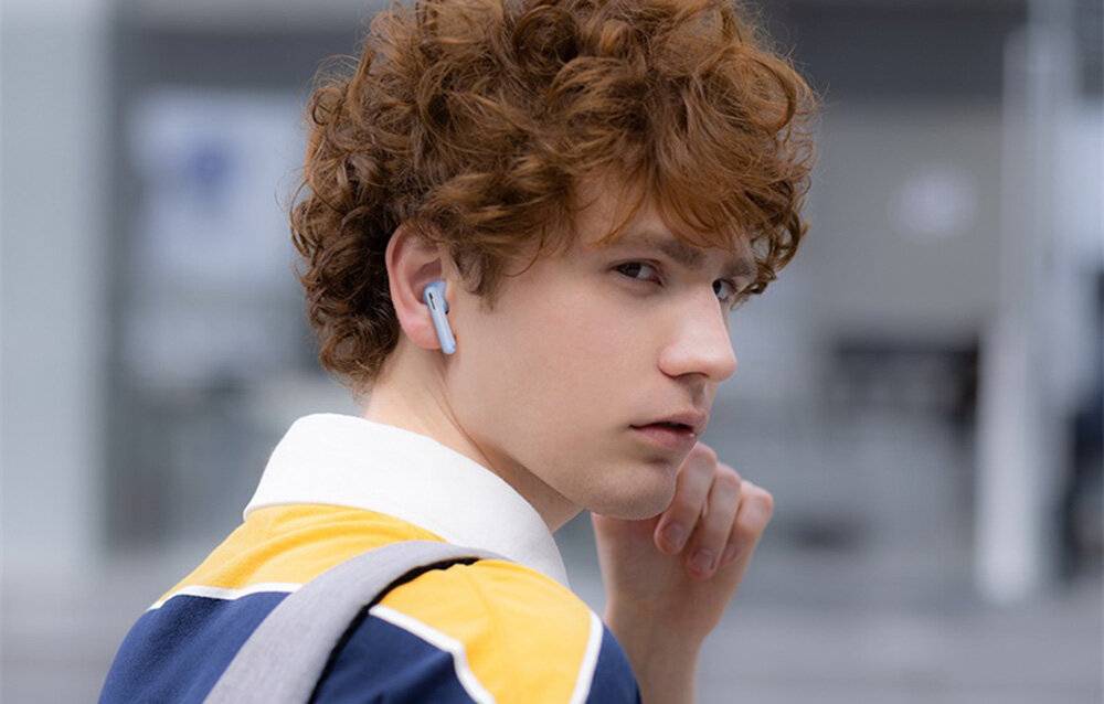 Eine Person mit lockigem Haar, die kabellose Ohrhörer trägt, schaut nachdenklich zur Seite mit einer Hand in der Nähe ihres Gesichts.