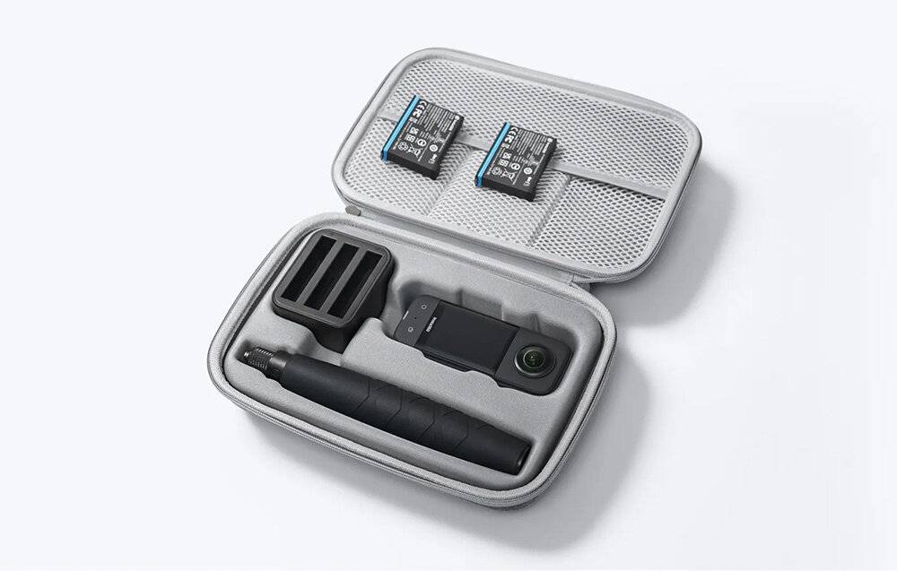 Eine kleine schwarze Kamera mit Objektiv, zwei Batterien und einem Griff sind ordentlich in einem offenen grauen Koffer auf weißem Hintergrund angeordnet.