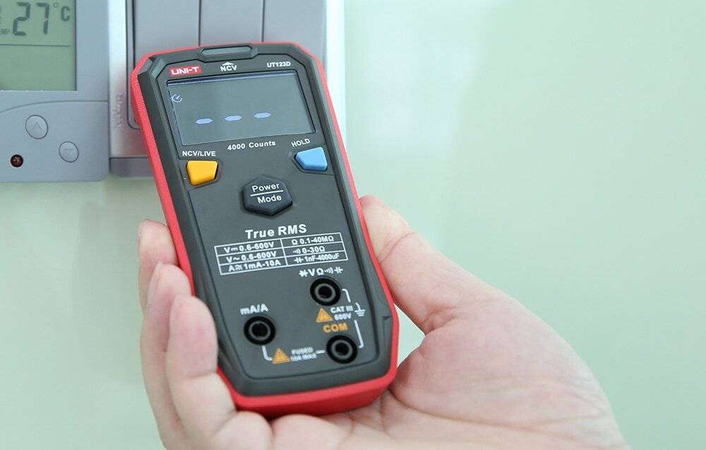 Eine Person hält ein rot-schwarzes digitales Multimeter, das Zahlen anzeigt. Das Multimeter wird zur Messung verschiedener elektrischer Größen verwendet.