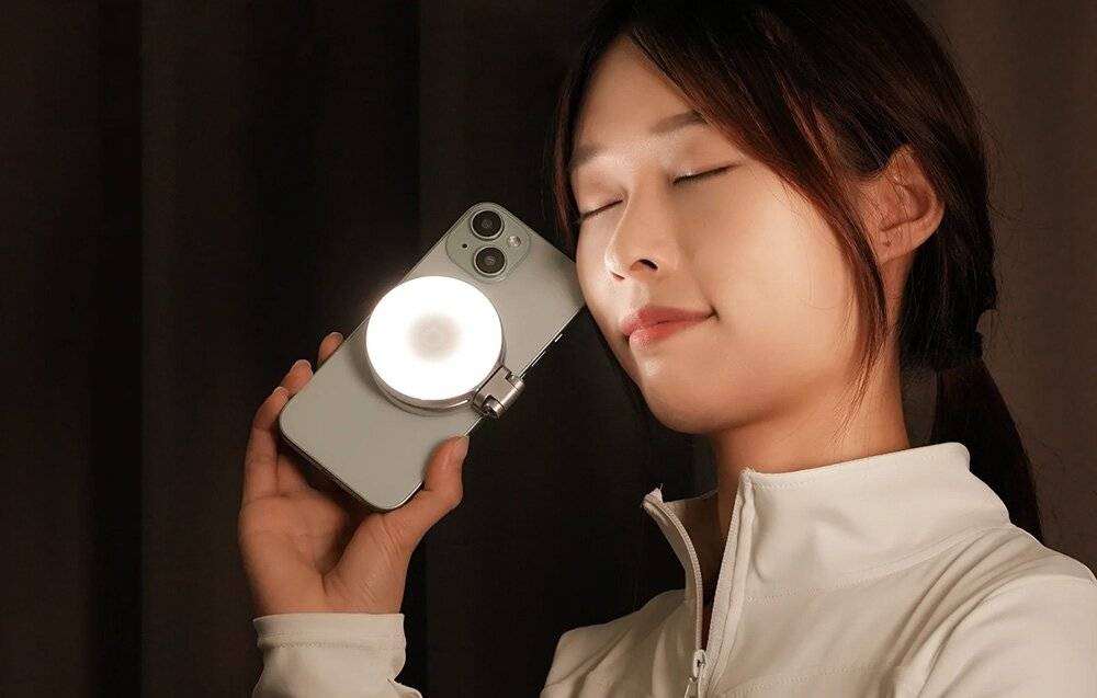 Eine Person, die ein Smartphone mit einer daran befestigten runden Leuchte hält, mit einem entspannten Gesichtsausdruck während einer Fotosession posiert.