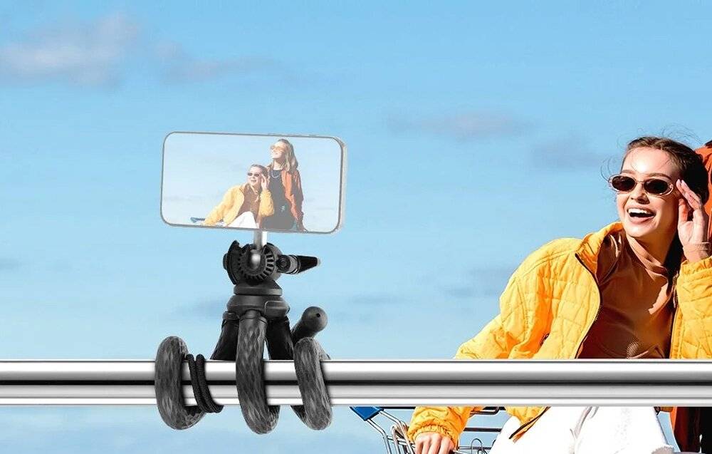 Eine Person in einer gelben Jacke macht ein Selfie mit einem Telefon, das auf einem flexiblen Stativ montiert ist, das um eine Metallstange im Freien gewickelt ist.
