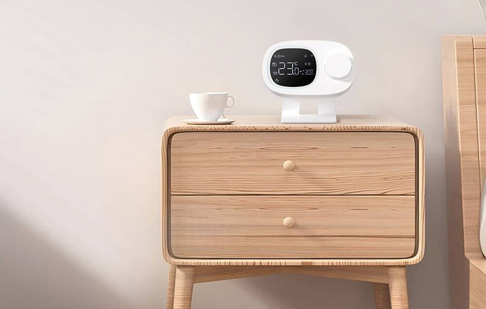 Ein hölzernes Nachttischchen mit einer modernen weißen digitalen Uhr, die '20:36' anzeigt, und Wettersymbolen. Eine weiße Tasse sitzt neben der Uhr.