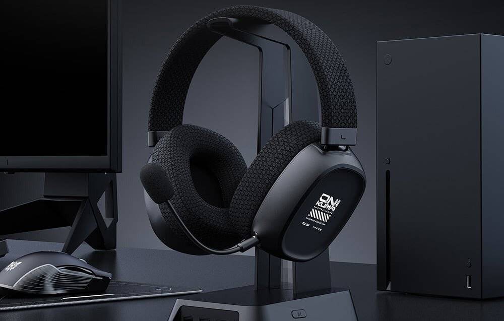Schwarzes Gaming-Headset mit gepolsterten Ohrmuscheln und angebrachtem Mikrofon, das auf einem schlanken Ständer neben einem Computermonitor ruht.