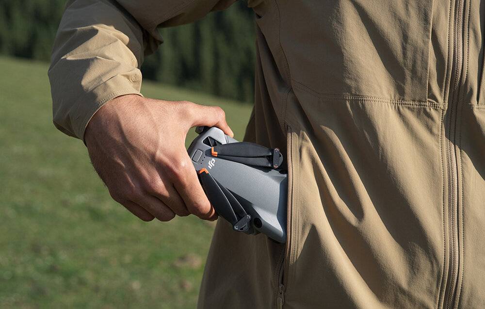 Eine Person in einer beigefarbenen Jacke hält eine zusammengeklappte Drohne und bereitet sich auf den Start in einer natürlichen Außenumgebung vor.