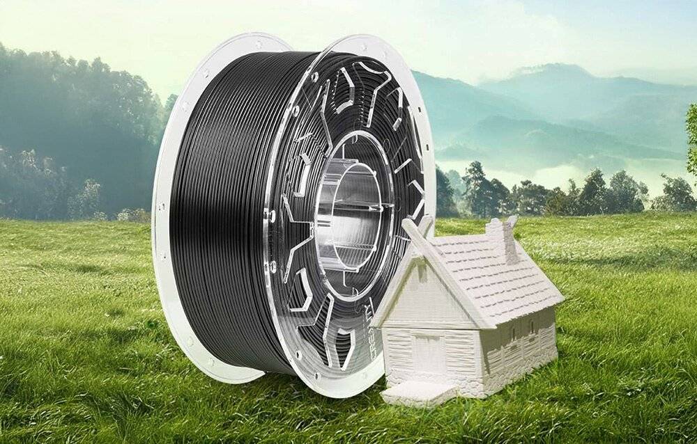 Eine große Spule schwarzes 3D-Druckfilament steht auf Gras neben einem kleinen weißen Modellhaus, mit Hügeln und Bäumen im Hintergrund.