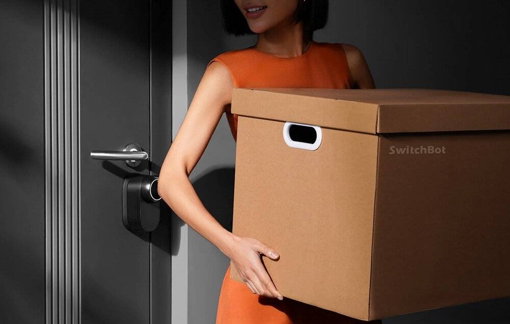 Eine Person in einem orangefarbenen Kleid trägt eine große Kartonschachtel durch eine Tür.