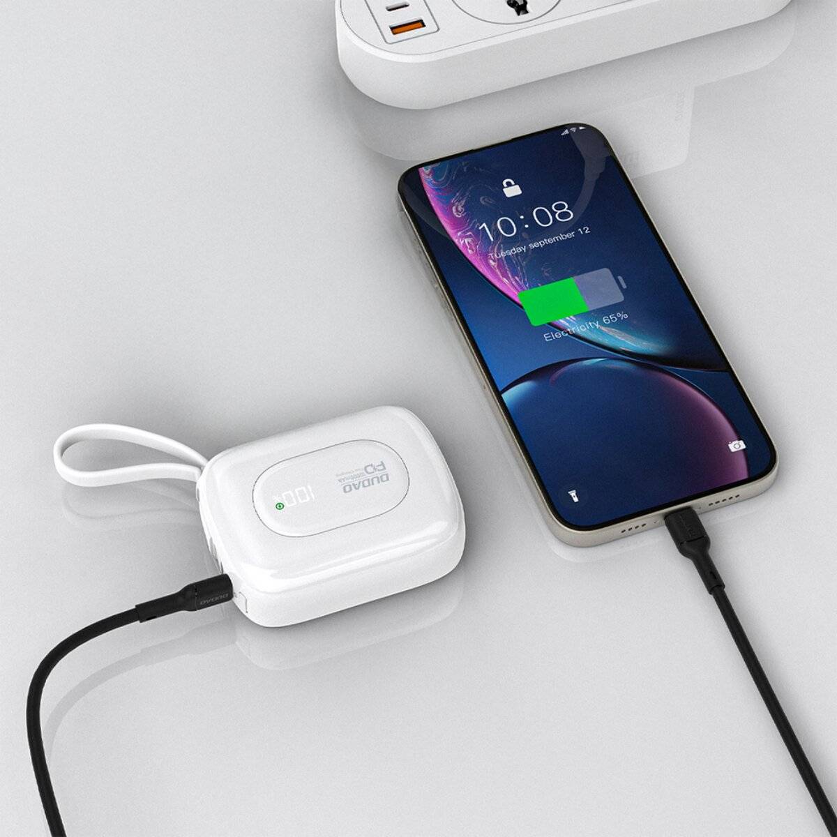 Ein Smartphone lädt drahtlos auf einem weißen Pad, das per Kabel verbunden ist. Eine Steckdose mit zwei sich aufladenden Geräten ist im Hintergrund zu sehen.