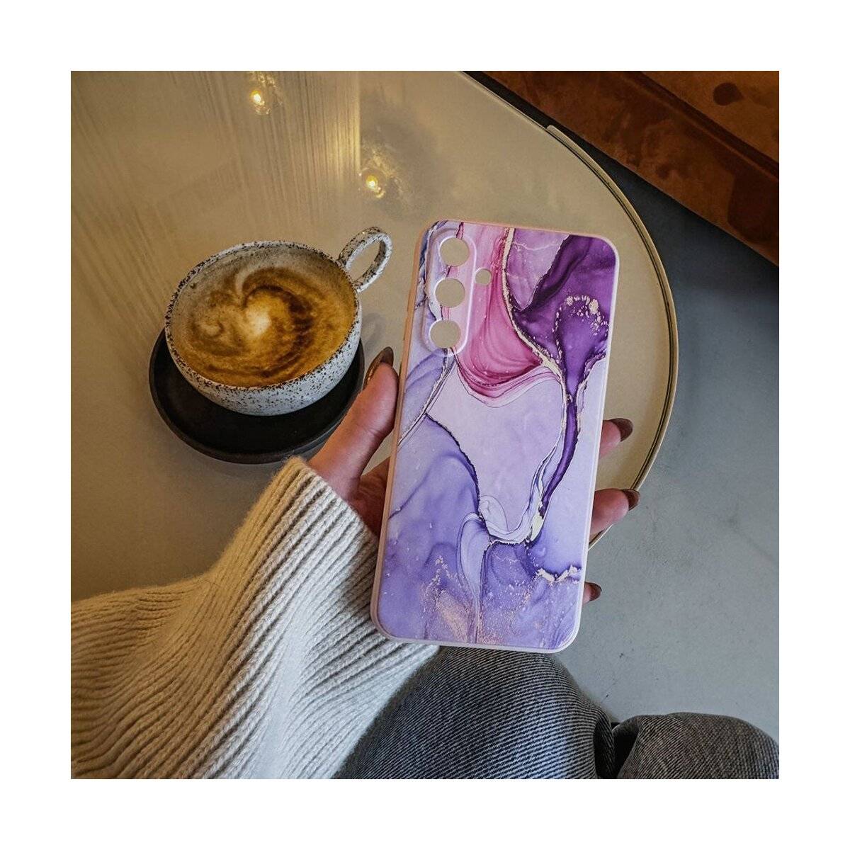 Eine Person hält ein Smartphone mit einer violetten und rosa abstrakten Design-Hülle über einem Tisch mit einer Tasse Cappuccino.