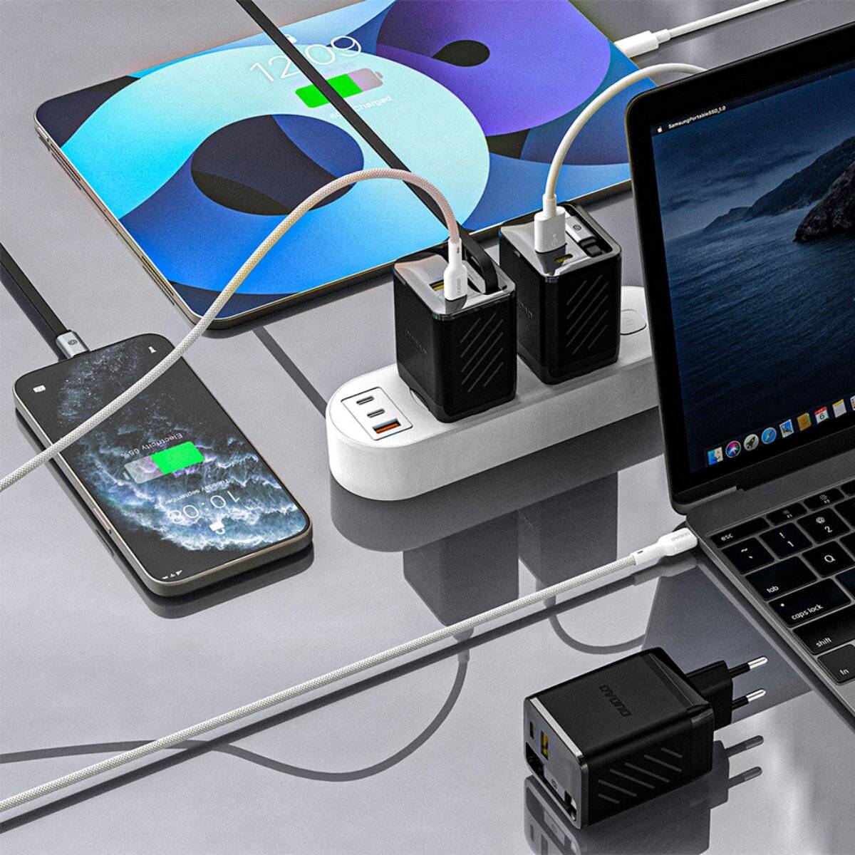 Mehrere Geräte, darunter ein Telefon und ein Laptop, werden auf einem Schreibtisch mit verschiedenen Kabeln und Adaptern aufgeladen, die in Steckdosenleisten eingesteckt sind.
