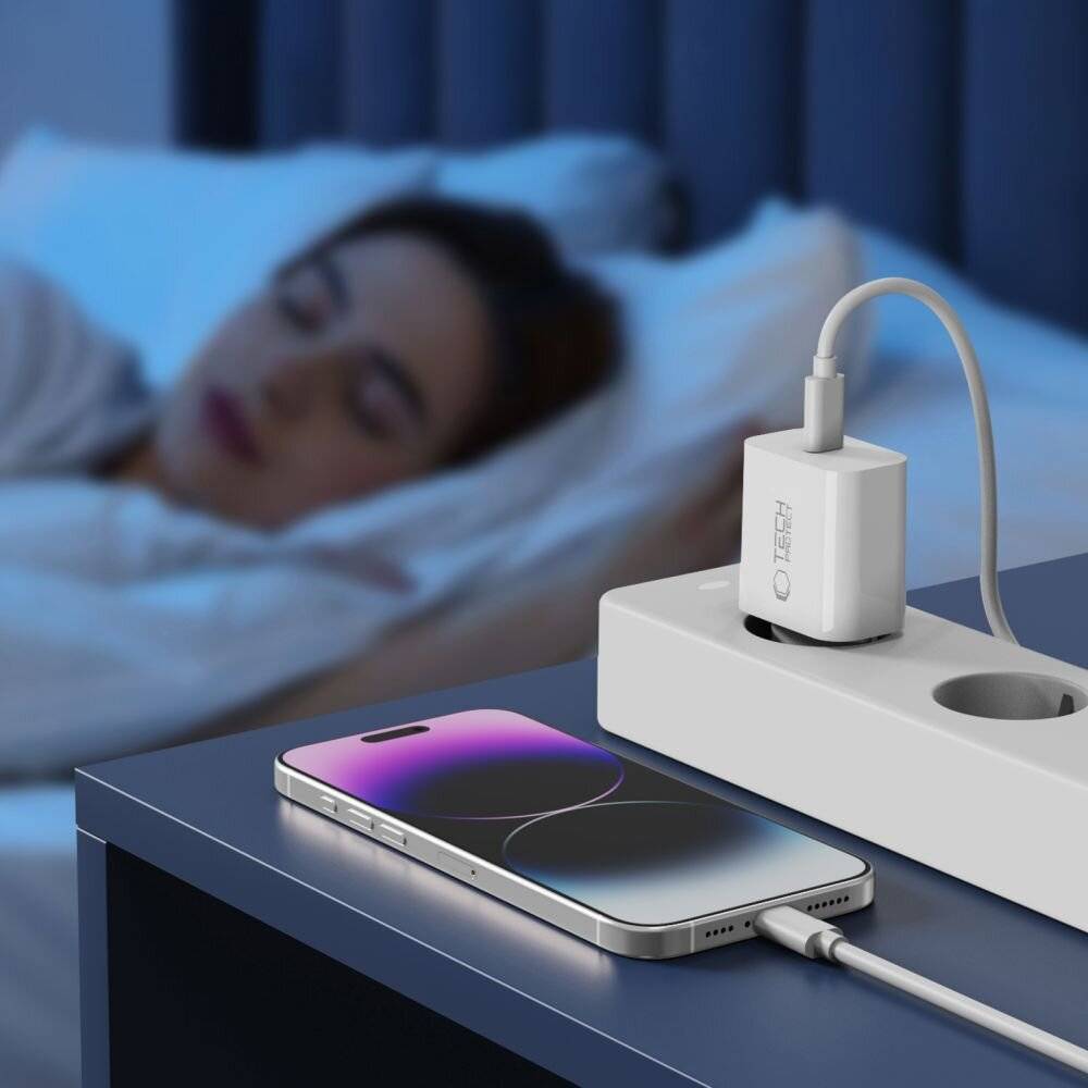 Ein Smartphone, das auf einer Steckdosenleiste neben einer schlafenden Person aufgeladen wird, wobei der Fokus auf dem Bildschirm des Telefons und der Ladeeinrichtung in einer ruhigen Umgebung liegt.