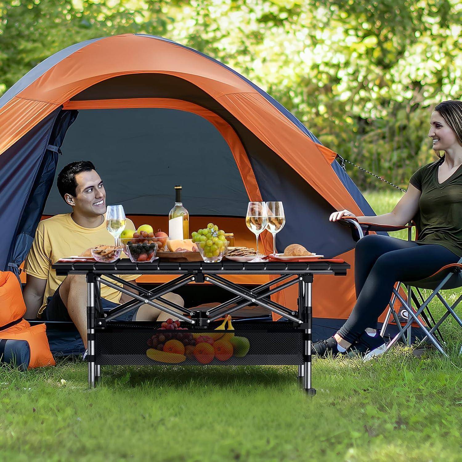 Ein Paar sitzt vor einem orangefarbenen Zelt und genießt ein Picknick mit Getränken und Früchten auf einem Tisch, umgeben von einer Grünfläche.