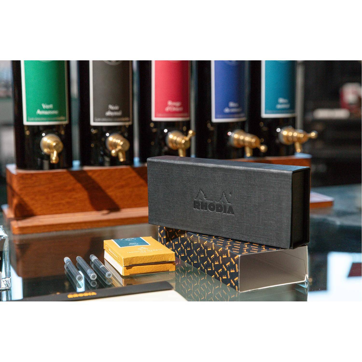 Ein schwarzes und orangefarbenes Schreibwarenset mit Rhodia-Branding, bestehend aus Stiften und Notizbüchern, die vor farbigen Getränkespendern ausgestellt sind.