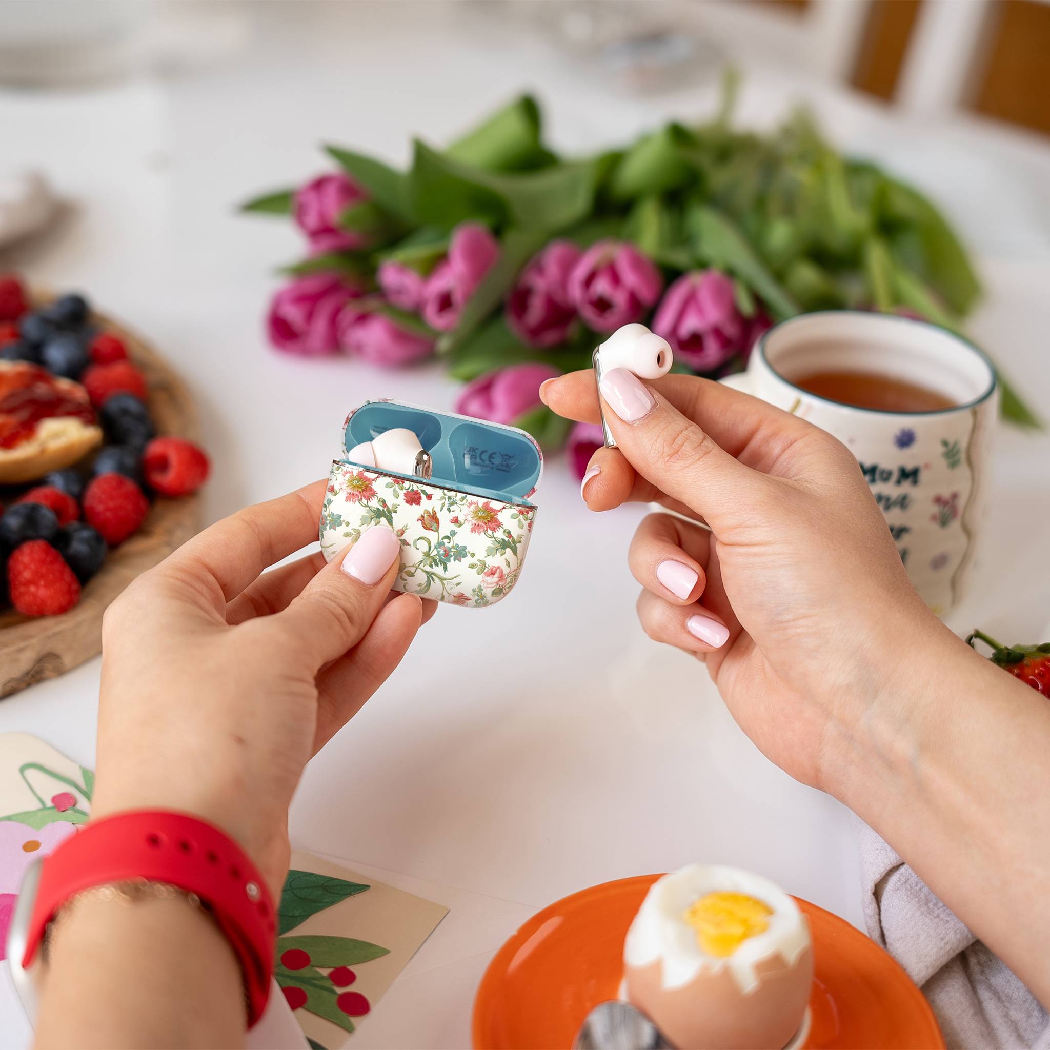 Eine Person hält ein Paar Ohrstöpsel in der Hand, eines in der Hand und das andere in einem Blumenetui. Ein Frühstückstisch mit Früchten, Tee und Blumen ist im Hintergrund zu sehen.