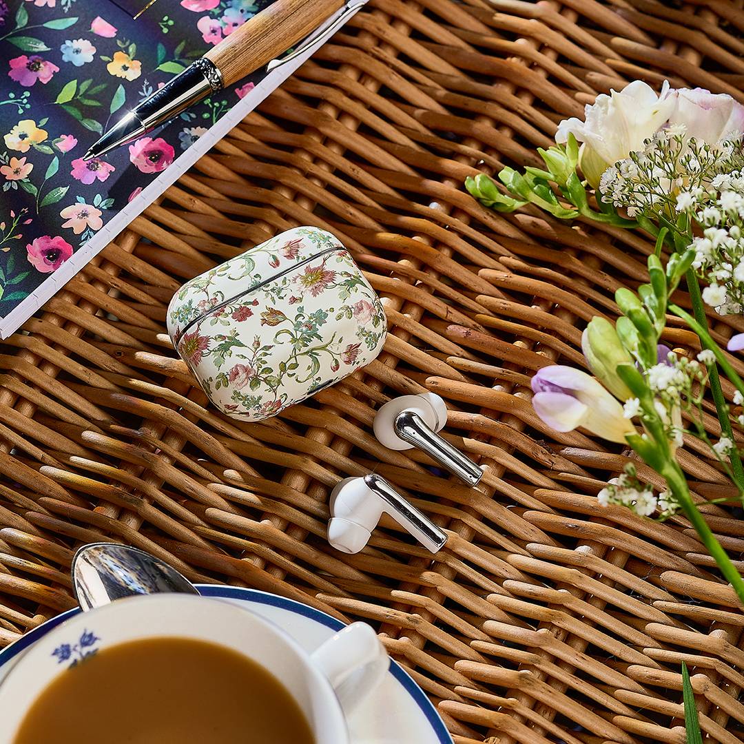 Ein florales Notizbuch und Stift, ein blumenverziertes Kopfhörertaschengehäuse mit Kopfhörern auf einer Wickeloberfläche, umgeben von frischen Blumen und einer Tasse Kaffee.