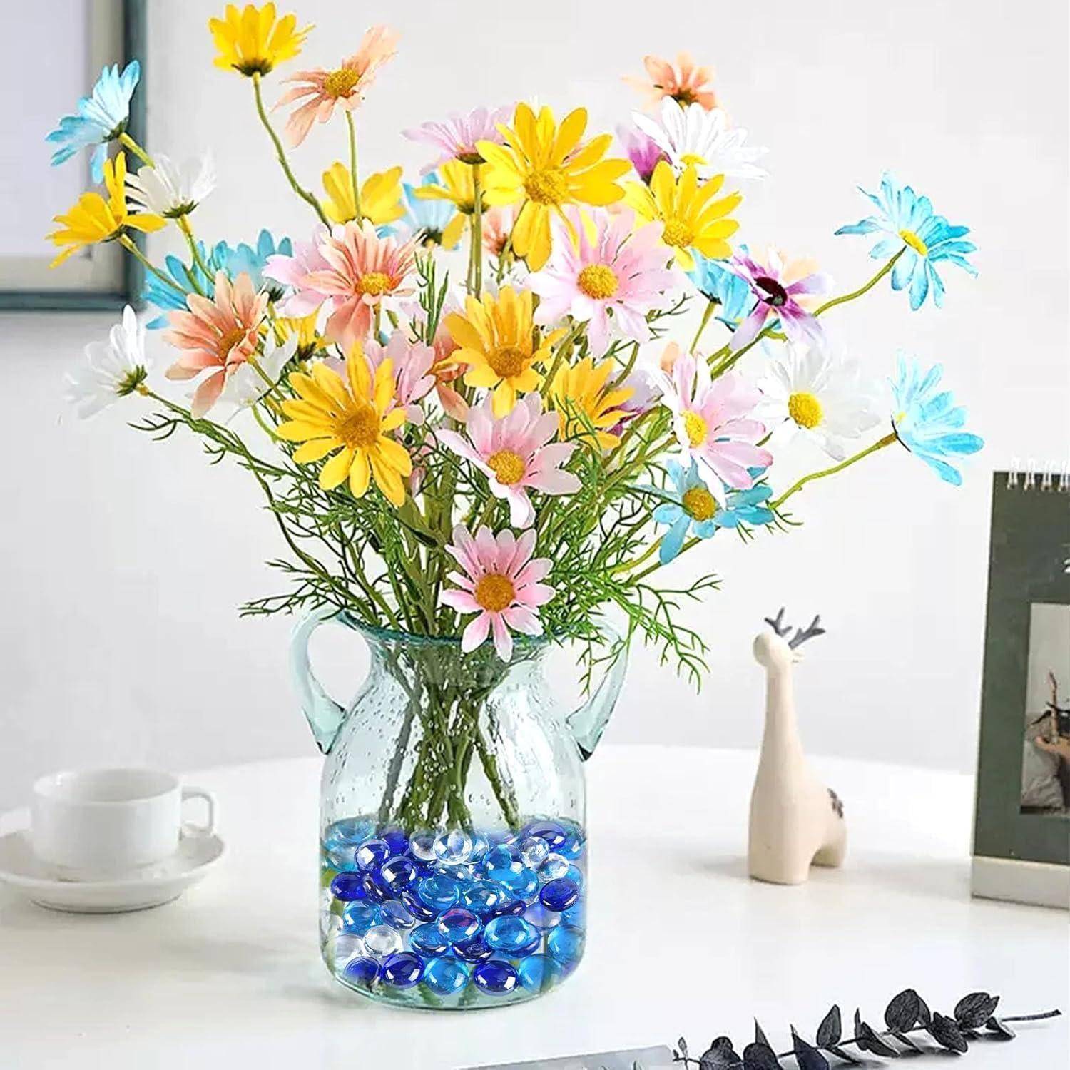 Eine Glasvase mit bunten Gänseblümchen und blauen Glaskieseln, auf einem weißen Tisch neben einer Kaffeetasse und einem Dekorationsobjekt gestellt.