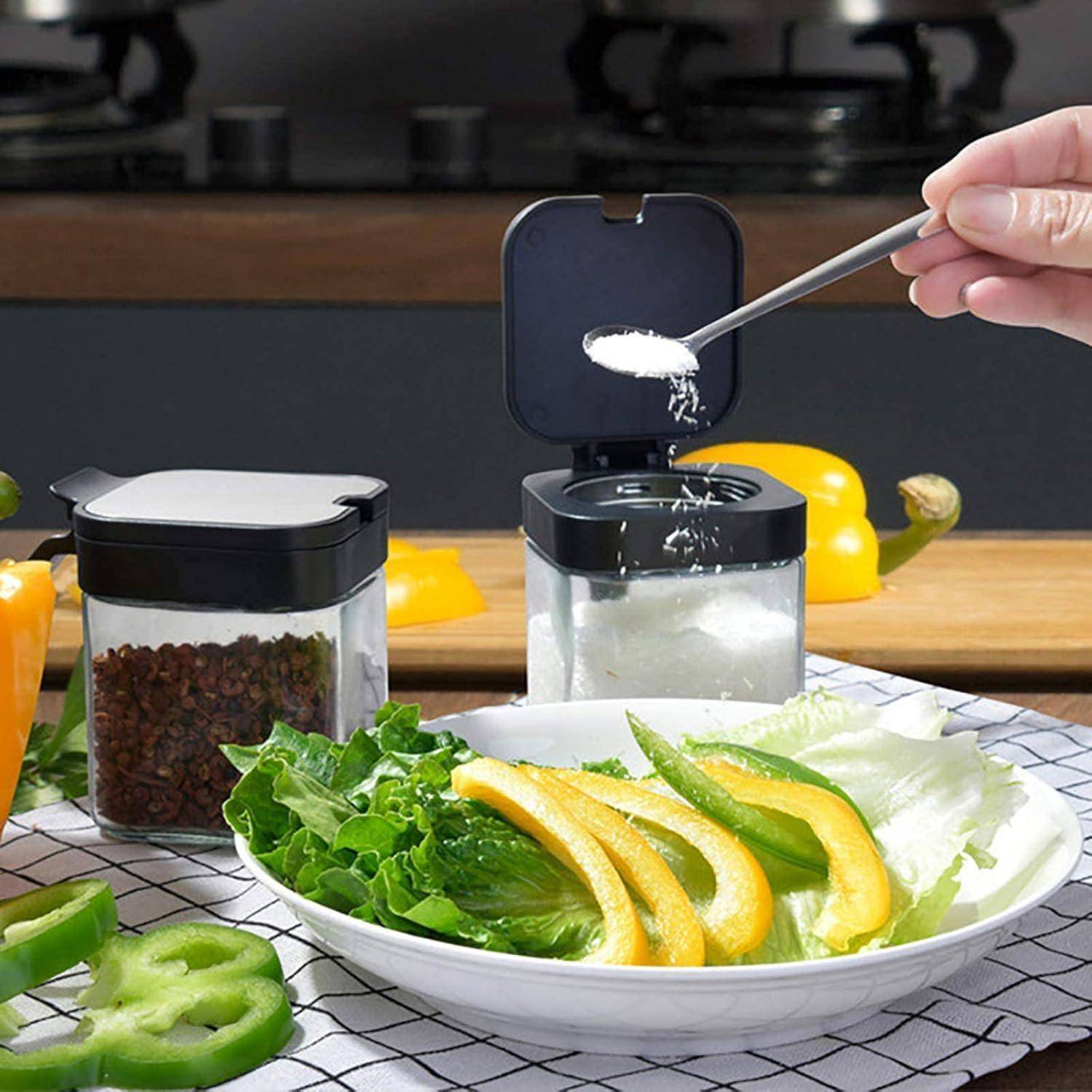 Eine Hand streut Salz über einen Teller mit Salat und gelben Paprikaschoten. In der Nähe befinden sich Behälter mit Pfeffer und Salz auf einer Küchentheke.