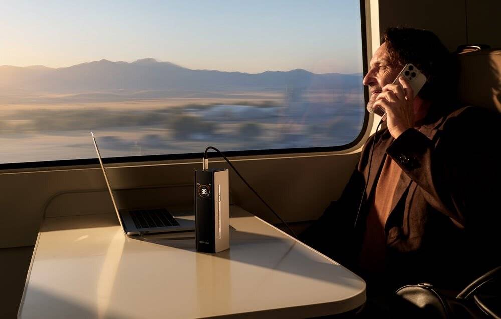 Eine Person spricht am Telefon, während sie in einem Zug sitzt. Ein Laptop und ein tragbares Gerät befinden sich auf einem Tisch. Eine malerische Landschaft ist außerhalb sichtbar.