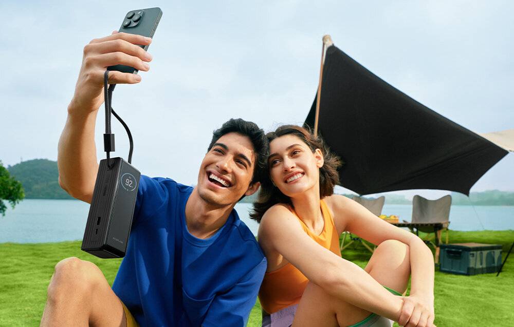 Ein lächelndes Paar macht ein Selfie im Freien und hält eine Powerbank in der Hand, die mit seinem Telefon verbunden ist. Im Hintergrund ist ein Zelt zu sehen.