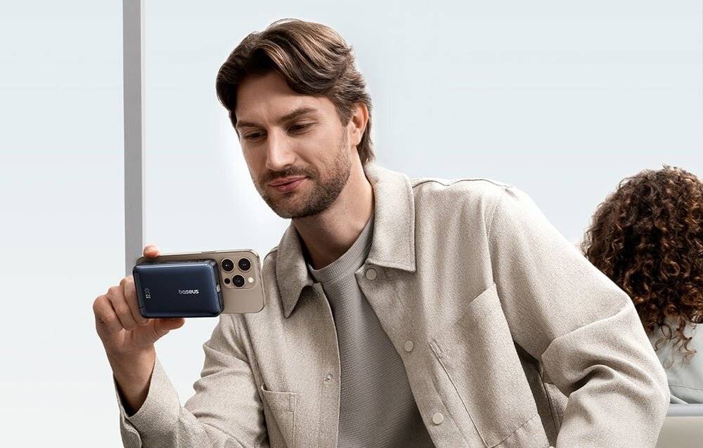 Eine Person hält ein goldenes Smartphone, das an eine Powerbank angeschlossen ist, und scheint etwas auf dem Bildschirm zu erfassen oder anzusehen. Die Umgebung ist im Inneren.