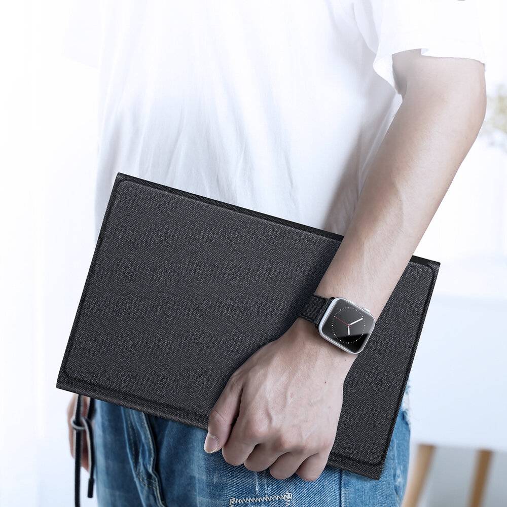 Eine Person trägt eine Smartwatch und hält ein geschlossenes schwarzes Notizbuch in der Hand, während sie vor einem hellen Hintergrund steht.