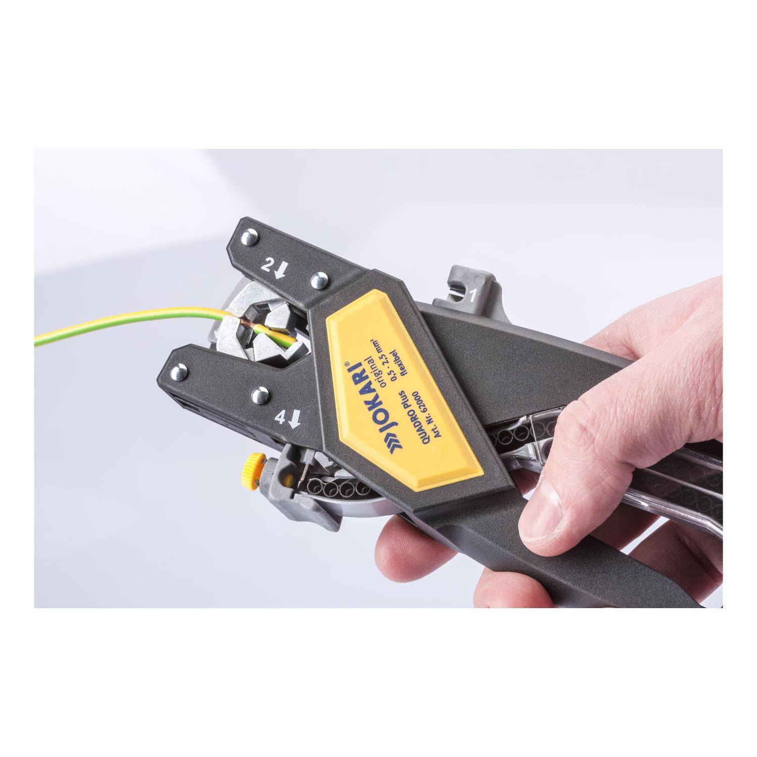 Eine Hand benutzt ein Jokari-Abisolierwerkzeug an einem grün und gelb gestreiften Elektrokabel und demonstriert dessen Schneid- und Abisolierfunktion.