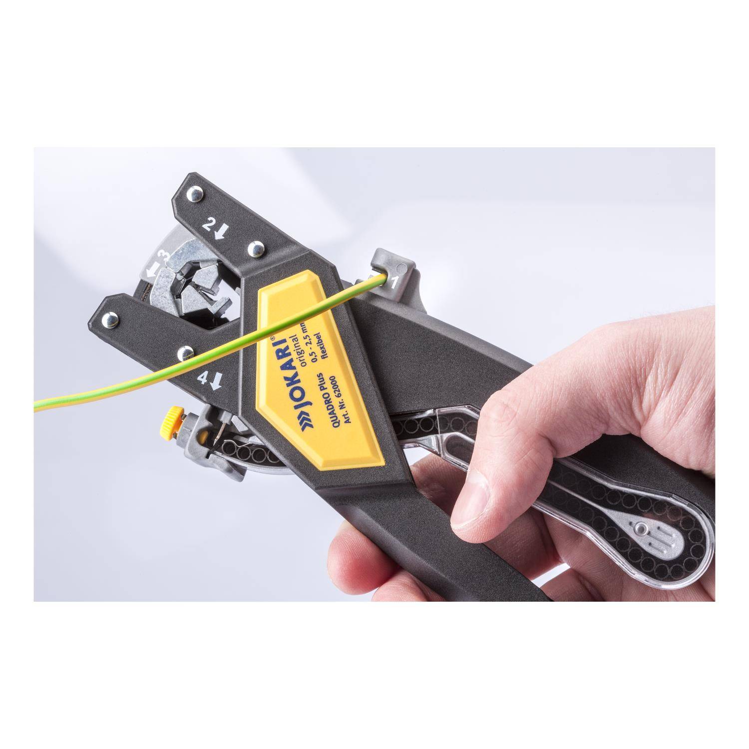 Eine Hand einer Person, die einen Jokari-Abisolierzange verwendet, um gelbe Isolierung von einem grünen Draht zu entfernen, wobei die Werkzeugbenutzung demonstriert wird.