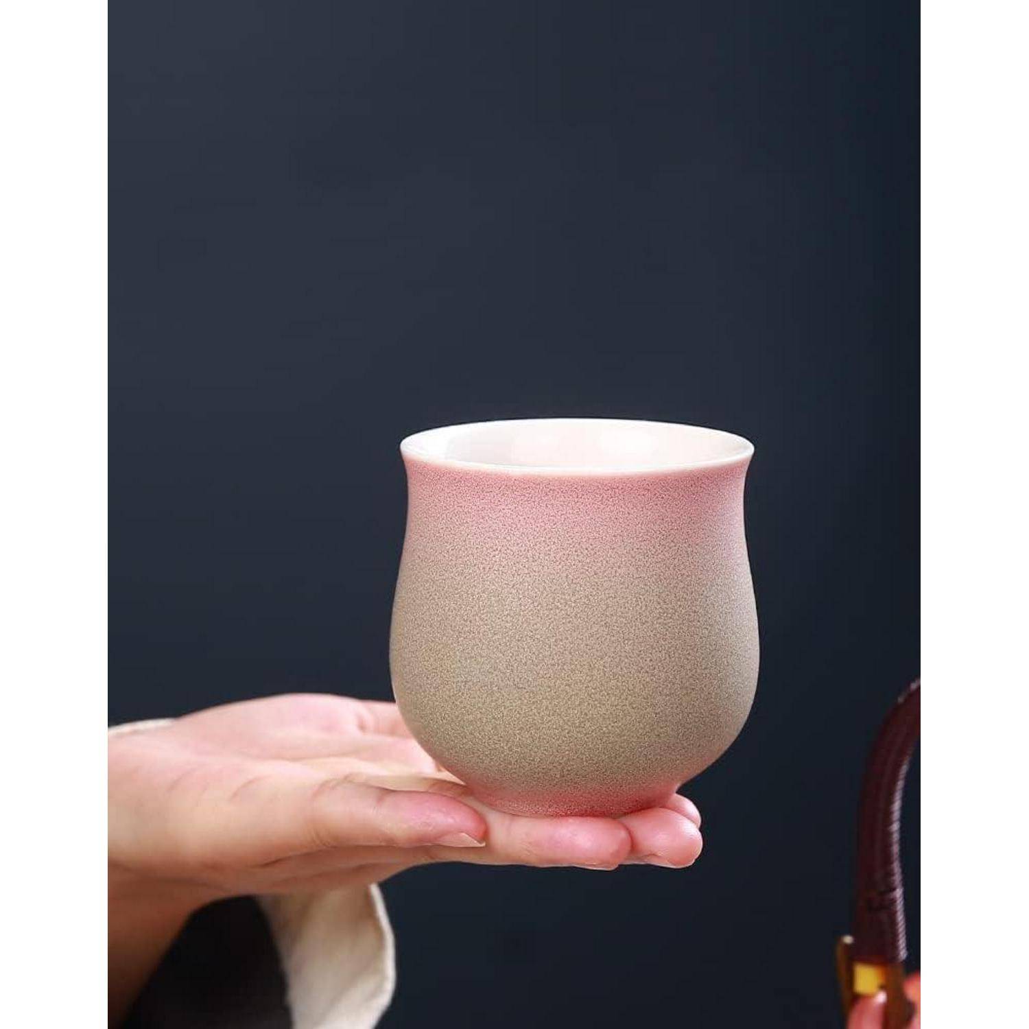 Eine Hand, die eine kleine, abgerundete Keramiktasse mit einem rosa- und grauem Farbverlauf vor einem dunklen Hintergrund hält.