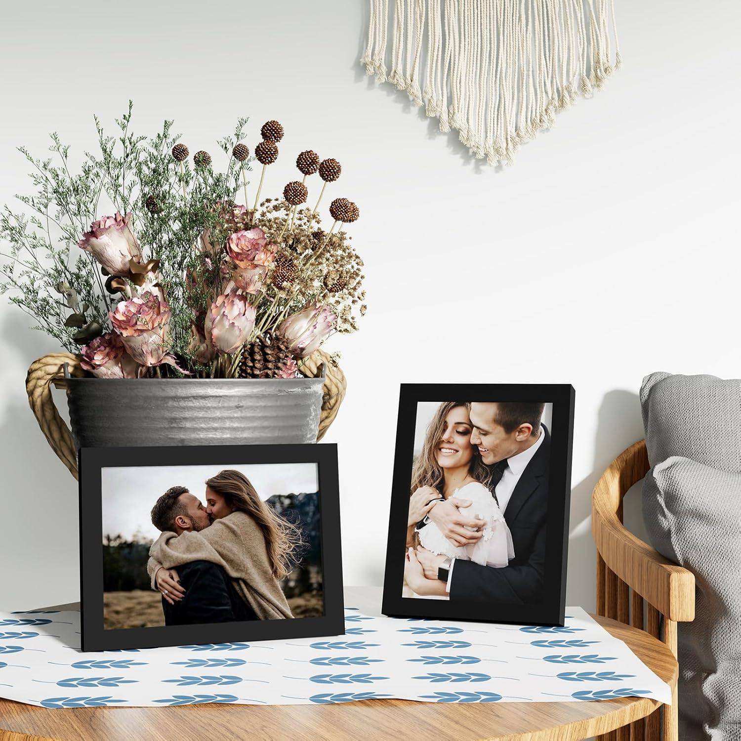 Zwei gerahmte Fotos auf einem Tisch: ein Paar, das sich umarmt, und ein anderes Paar, das ein Baby hält. Getrocknete Blumen in einer Vase, makramé-Wandbehang.